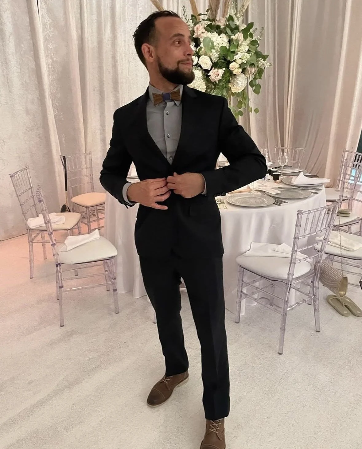 A man in a black suit adjusting his jacket at a formal event, standing in front of a decorated table with chairs and floral arrangements.