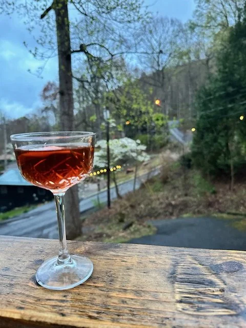 A glass of rosé wine on a wooden surface with a scenic view of trees, a road, and a cloudy sky in the background.