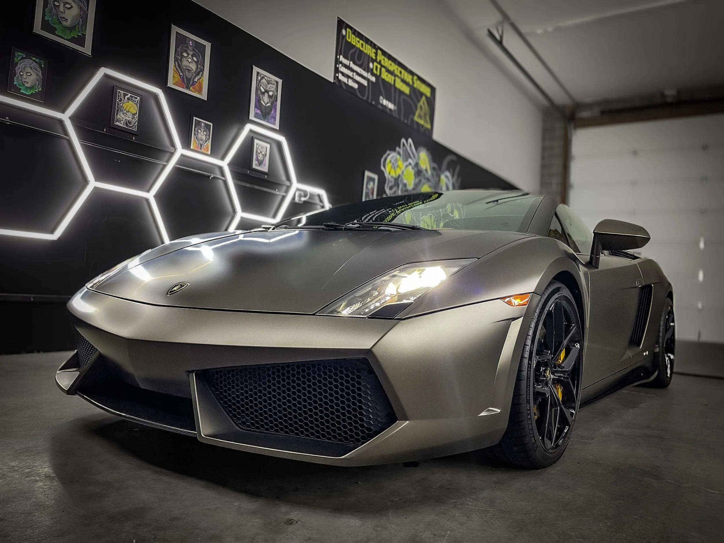 A metallic gray Lamborghini sports car parked in a garage with artistic wall decorations, including framed artwork and illuminated hexagonal shapes.