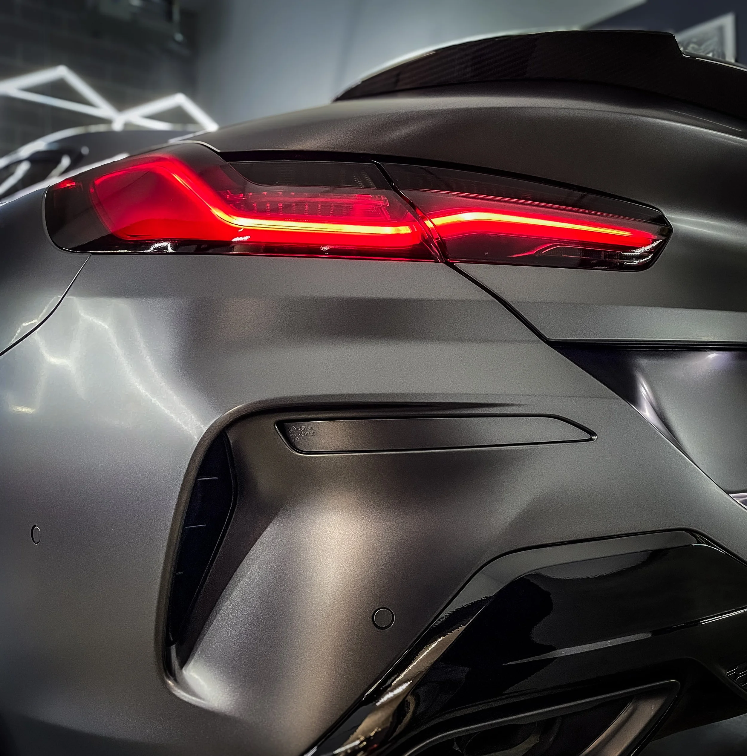 Close-up of the rear left side of a silver sports car, showing its illuminated red taillight and sleek design details.
