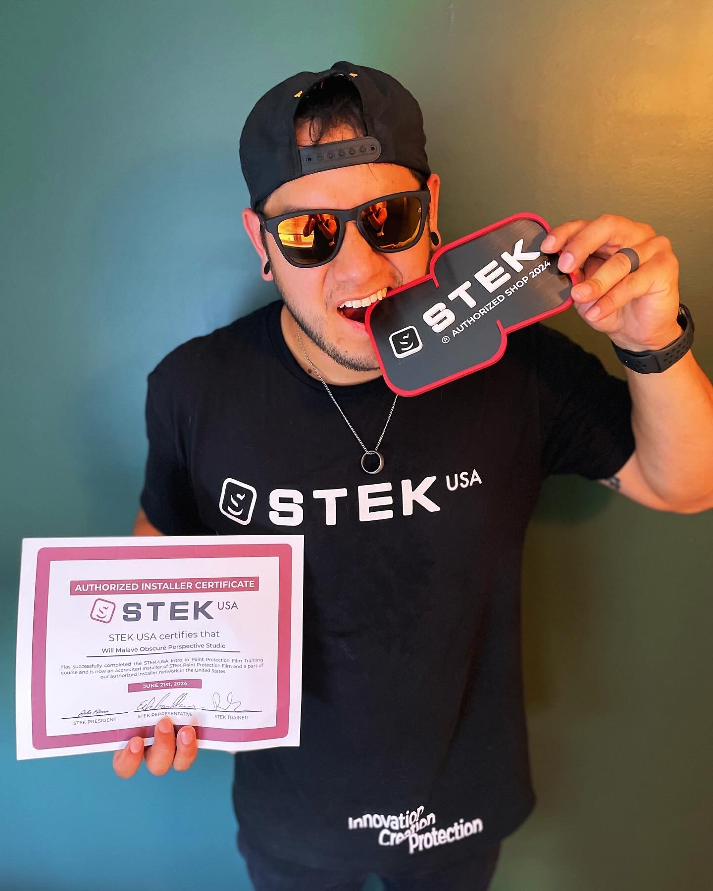 A man wearing sunglasses, a black cap backward, a black t-shirt with white text, and a watch is smiling and holding a sign that reads 'STEK Authorized Shop 2024.' He is also holding a certificate indicating he is an authorized installer for STEK USA. The background is a gradient of blue-green and yellow-orange.