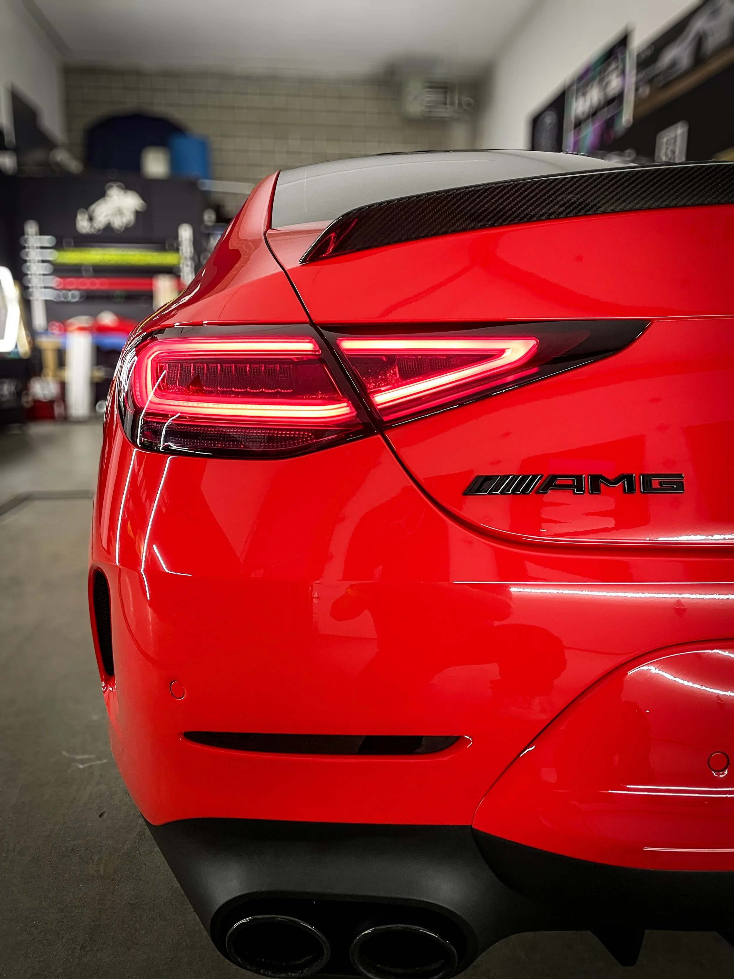 Close-up of the rear of a red Mercedes-AMG sports car, showcasing the taillight, AMG badge, and dual exhaust pipes, inside a garage or workshop.
