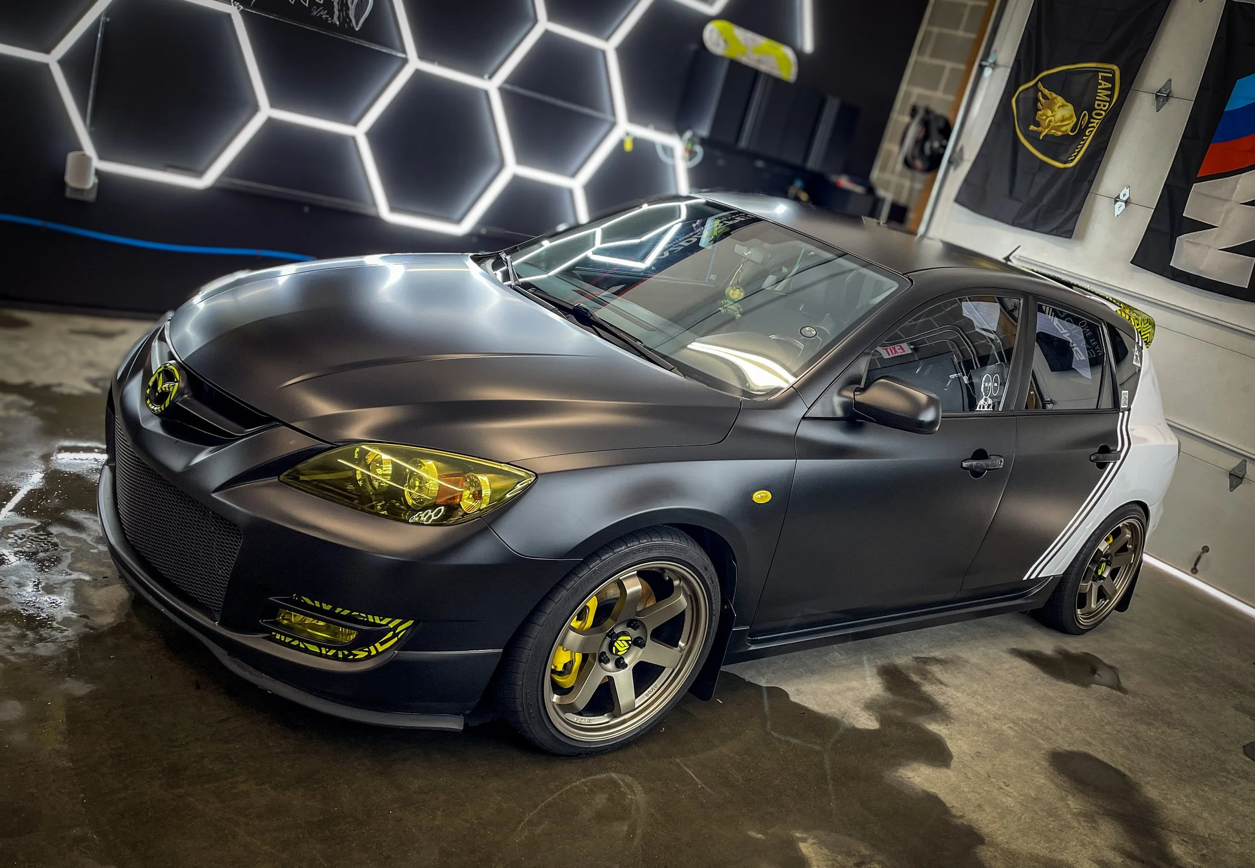 A black and silver sports car with yellow accents parked indoors, with a Lamborghini flag hanging on the wall and illuminated hexagon wall lights in the background.