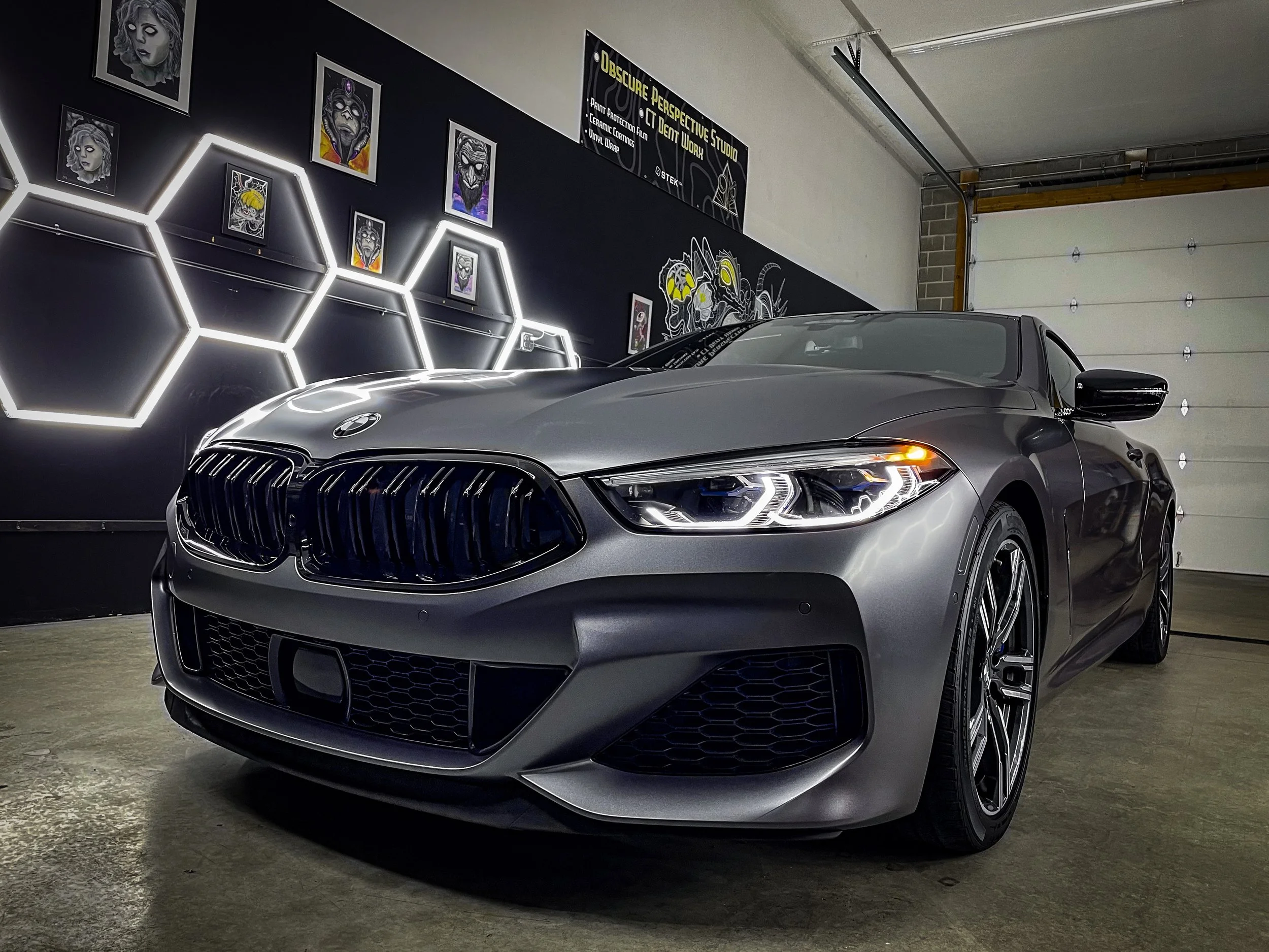 A silver BMW car parked in a garage with dark walls decorated with modern art and illuminated hexagonal light fixtures.