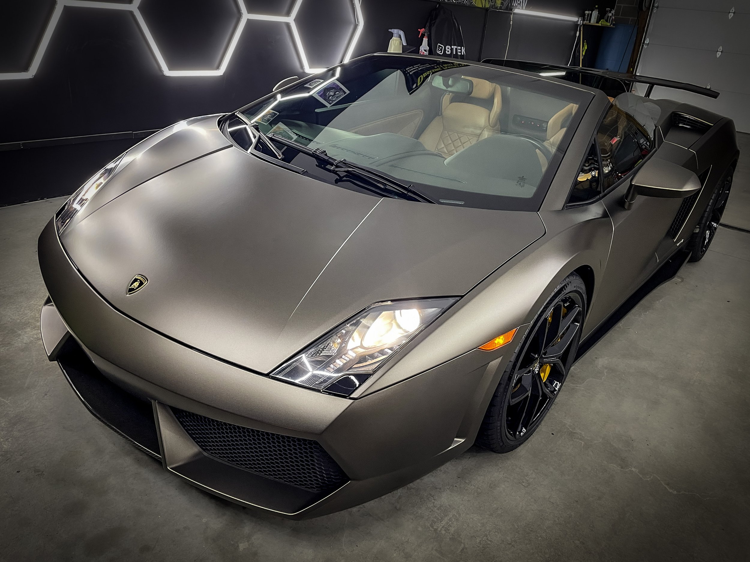 A matte grey Lamborghini Gallardo Spyder with black wheels and yellow brake calipers parked inside a garage with hexagonal wall lighting.
