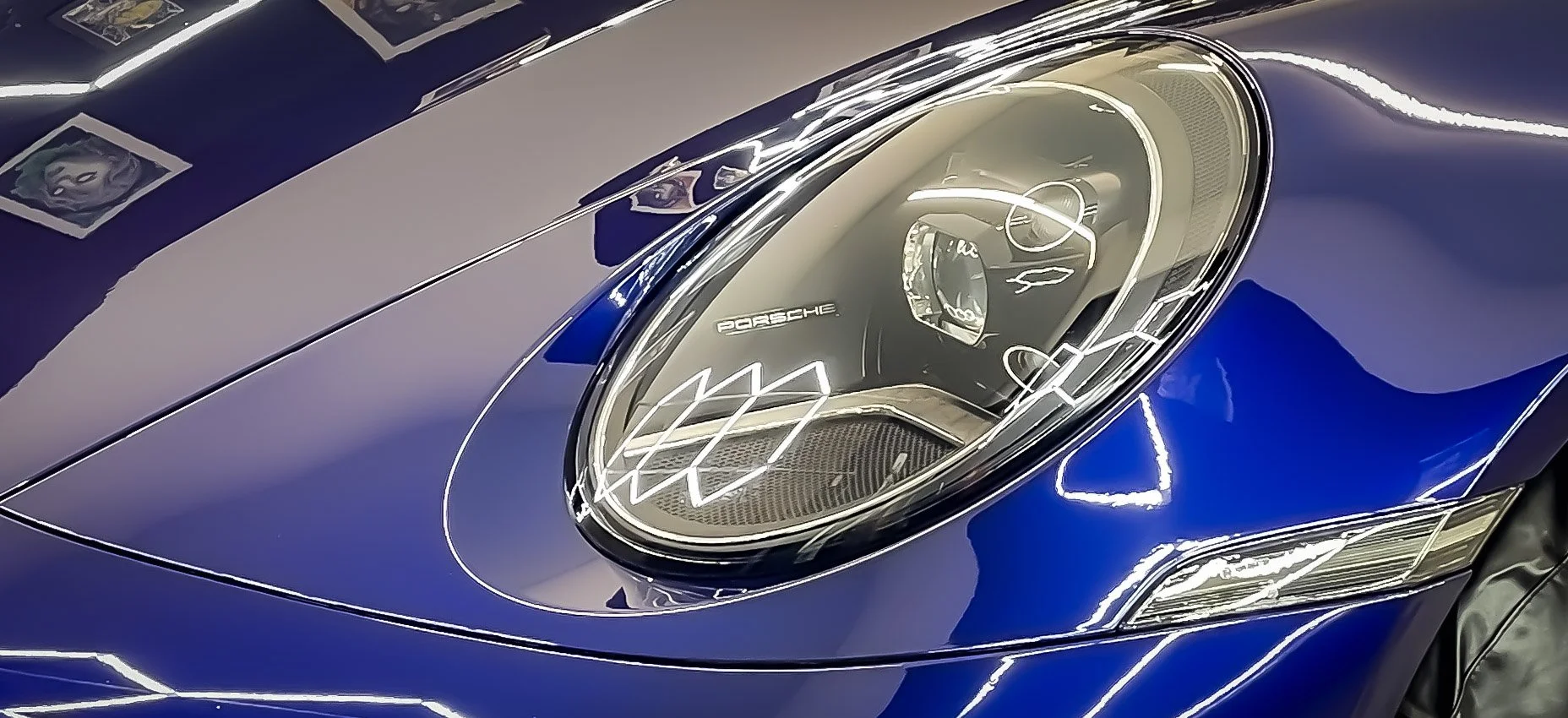 Close-up of a Porsche sports car headlight and front part, showing blue paint, intricate light design, and reflections.