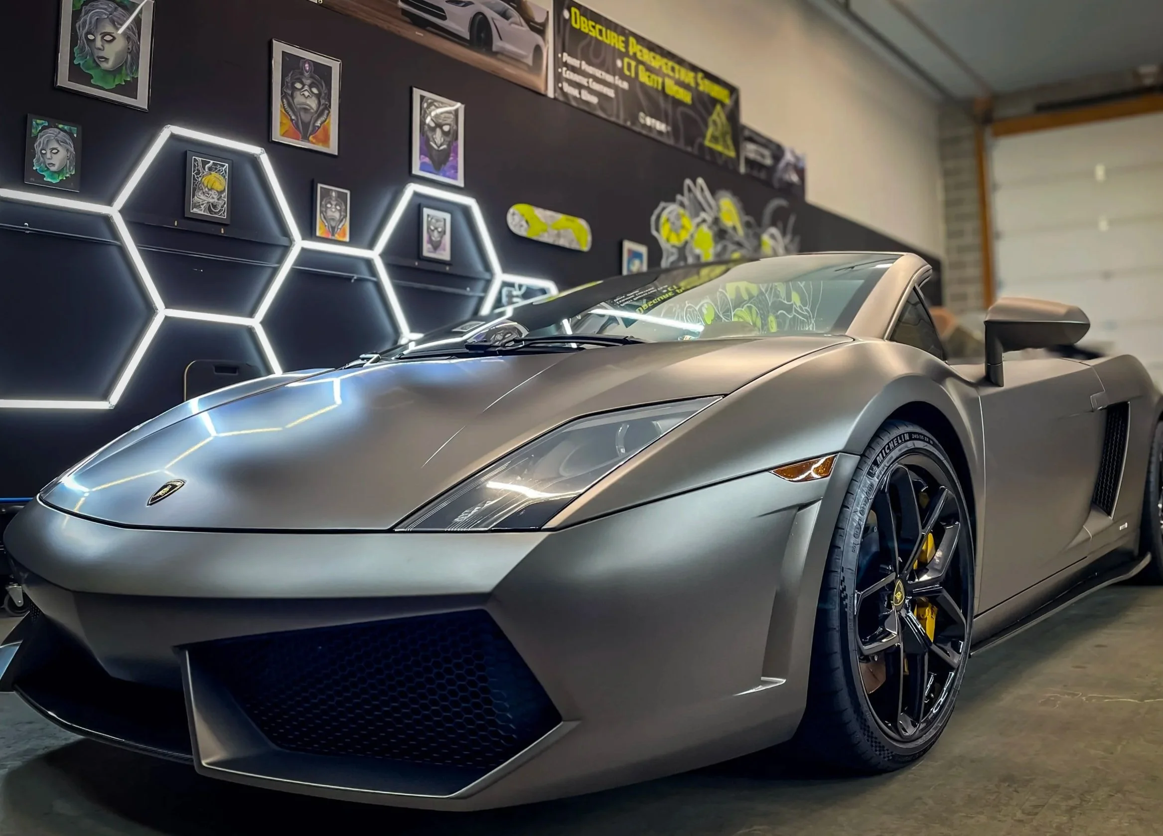 A silver Lamborghini sports car parked indoors in front of a black wall with framed artwork and a banner. The car has black wheels with yellow brake calipers and a sleek, aerodynamic design, that has been vinyl wrapped and ppf installed on front end