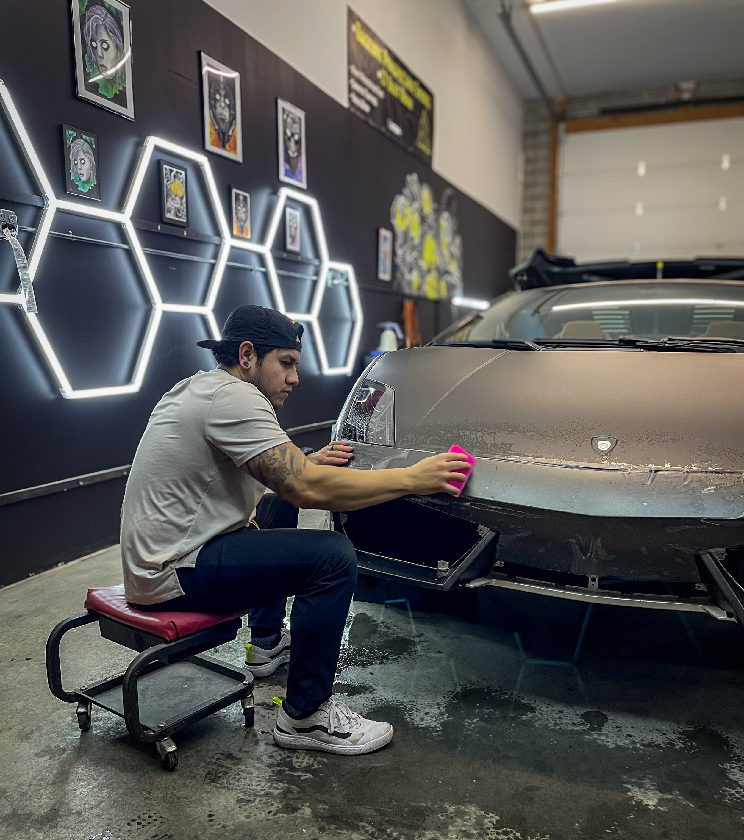 A man working on a car in a garage, seated on a rolling stool, wearing a beige t-shirt, black cap, and white sneakers, focused on the front of a silver car. The garage has black walls with illuminated hexagonal decorations and framed artwork. There's a blackboard with yellow writing on the wall, and a partially visible large unlabeled garage door.