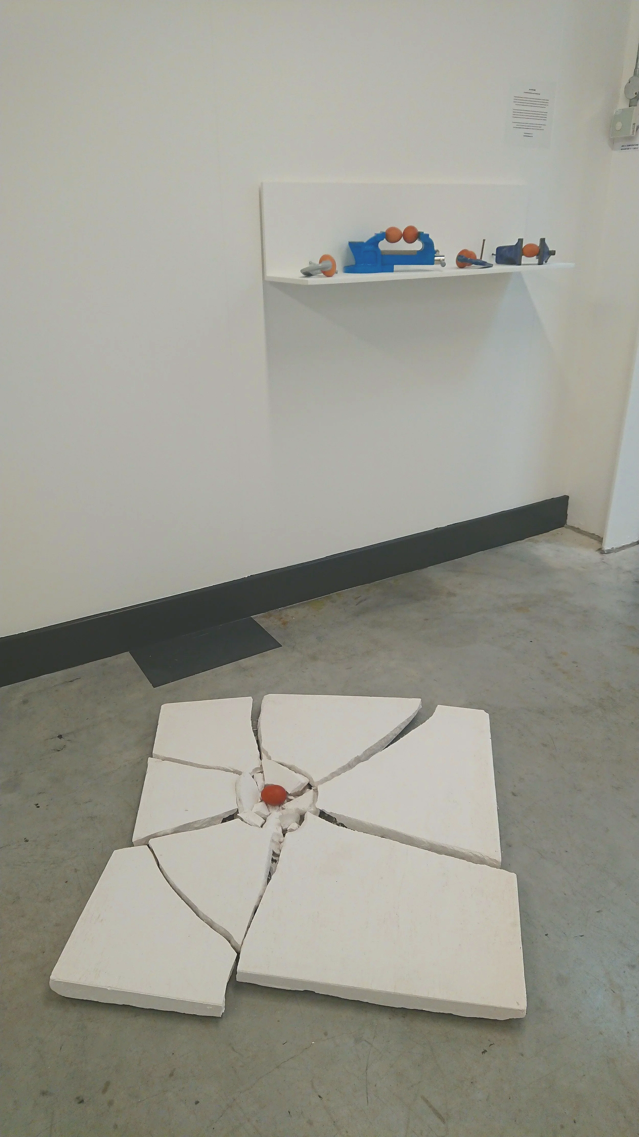 'Eggs in Vice' and 'Resilience' Shattered white ceramic tile with an egg in the center, on a gallery floor in front of a shelf holding several vice about to crush eggs.