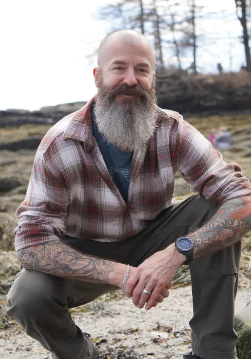 A bearded man with shaved head wearing a plaid shirt and watch, sitting outdoors on rocky ground with a forest background.