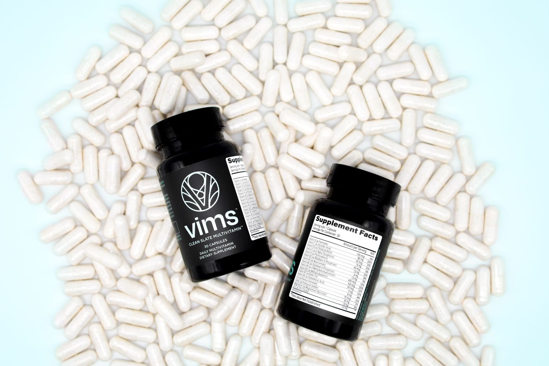 Two black supplement bottles surrounded by numerous white capsule pills on a light blue background.
