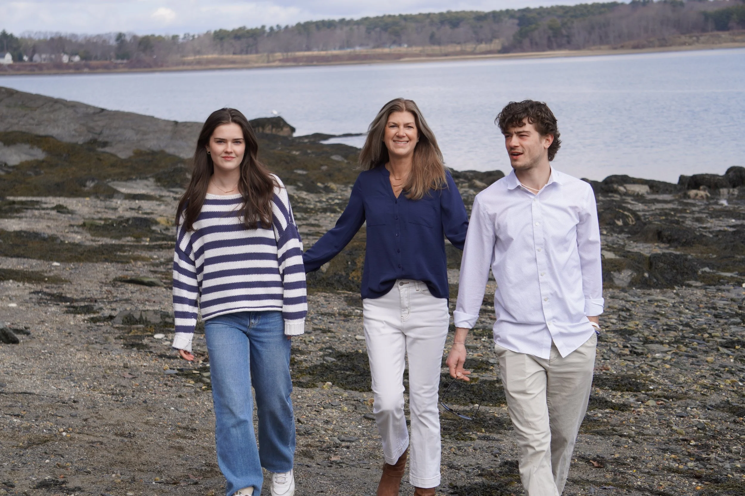 Three people walking along a rocky shoreline near a body of water, with a tree-lined hill in the background. The woman on the left has long brown hair and is wearing a striped sweater, the woman in the middle has long gray hair and is dressed in a dark blue shirt, and the man on the right has curly brown hair and is wearing a white shirt. They are smiling and talking.