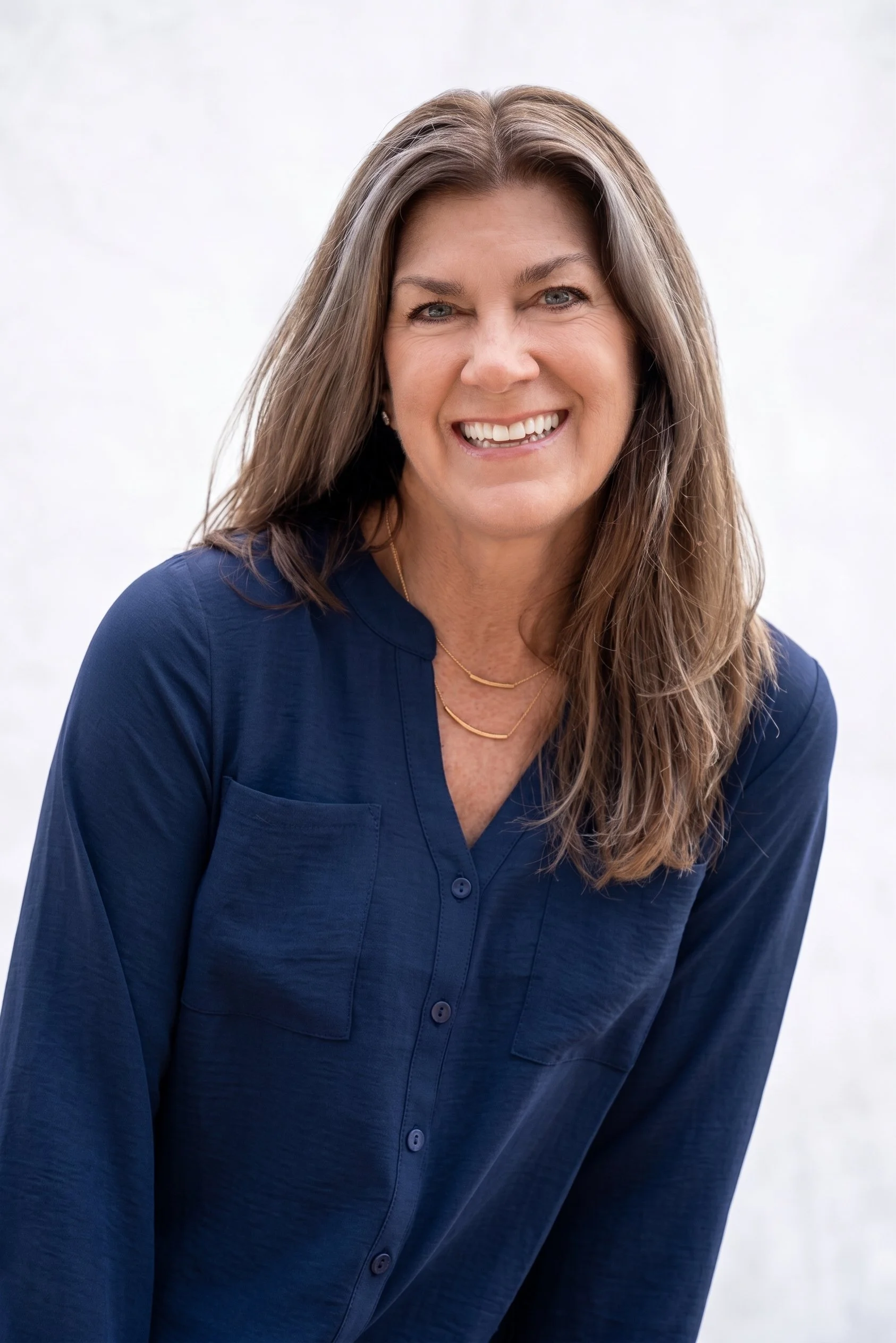 A woman with long brown hair smiling and wearing a navy blue button-up shirt and gold necklaces against a plain white background.