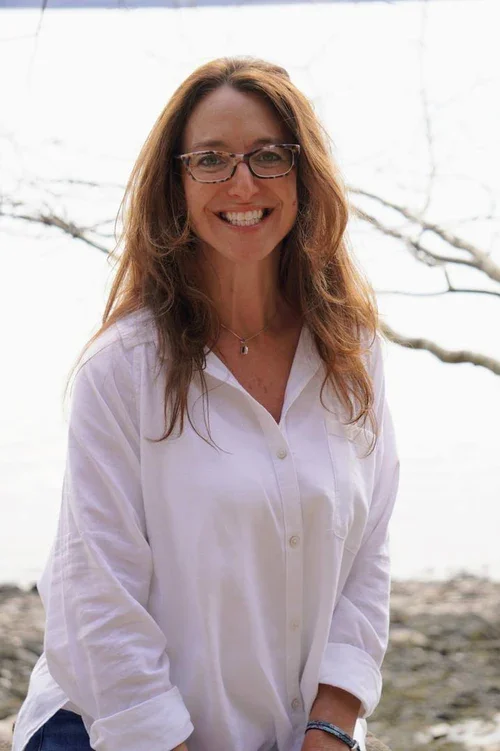 A woman with long wavy brown hair, glasses, and a white blouse smiling outdoors near a body of water.
