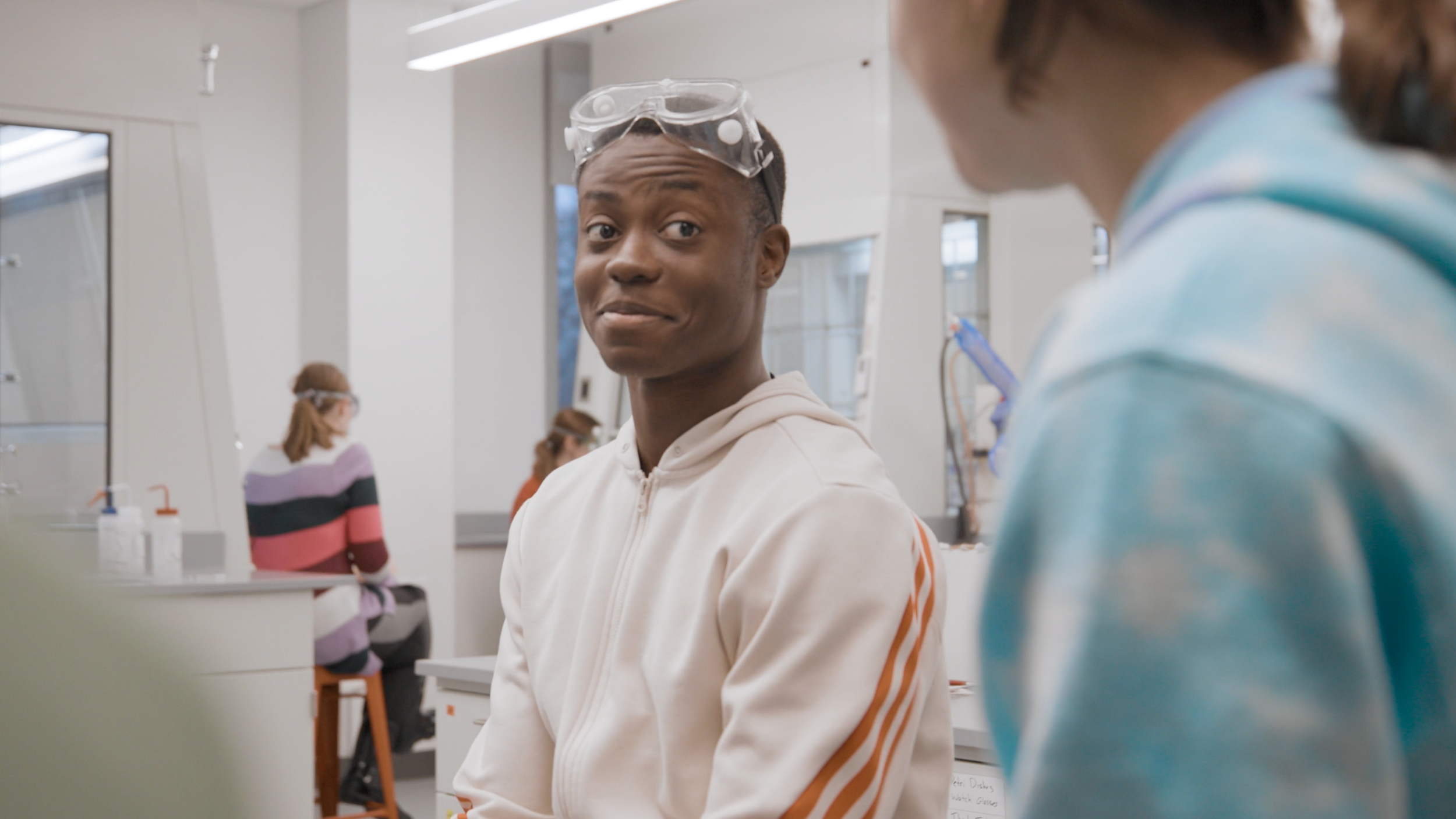 A young man with a curious expression wears safety goggles and a white hoodie with orange stripes as he looks at someone or something off-camera in a science lab.