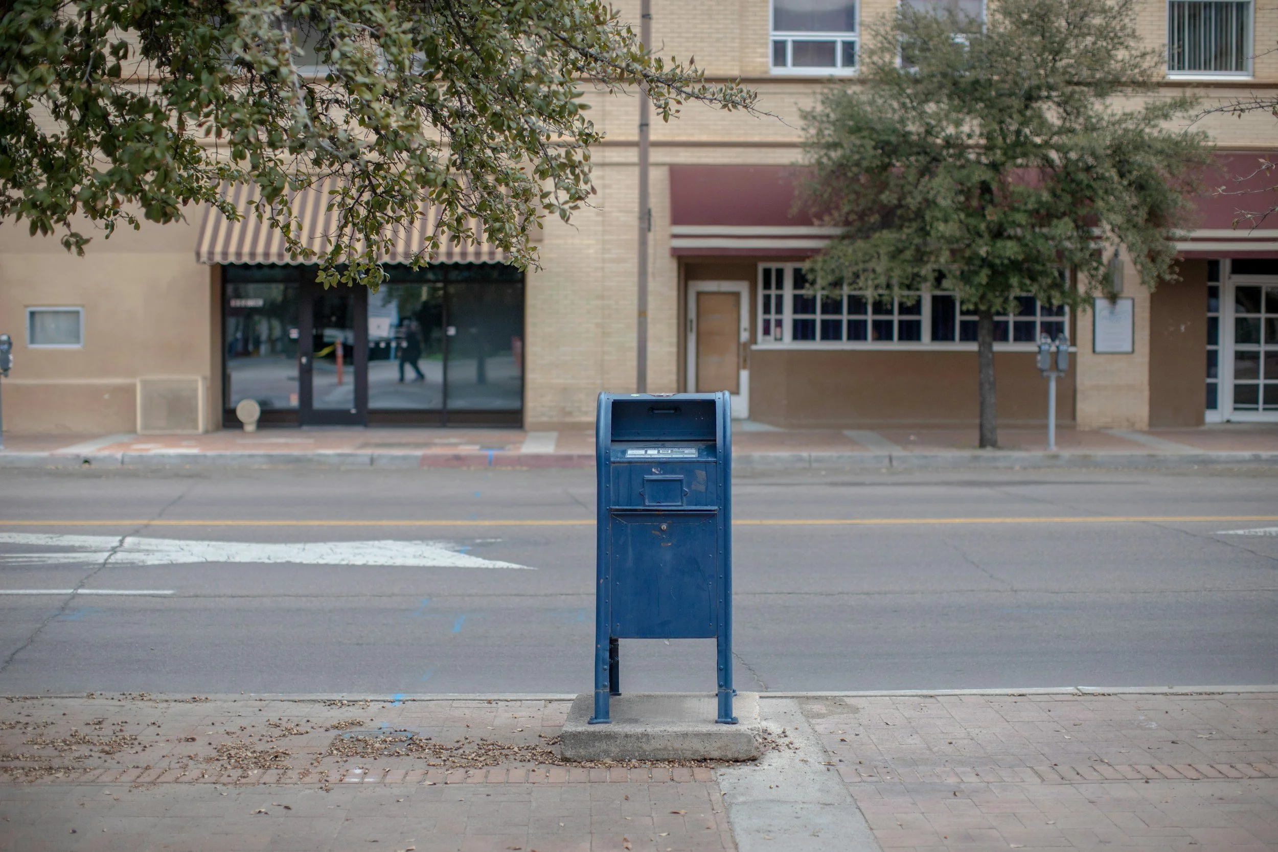 A blue mailbox is positioned in front of a sidewalk on a street. Behind the mailbox, there is a building with large windows, a door, and two trees with green leaves. The building has a beige brick facade with red awnings over the windows. There are a few parked cars reflected in the window and a person walking on the sidewalk.