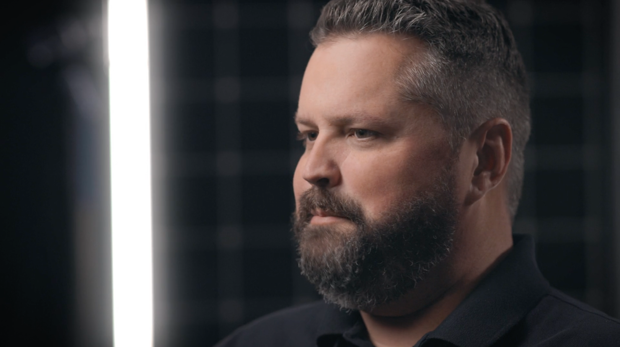 A man with a beard and short hair looking contemplative, with a bright vertical light behind him.