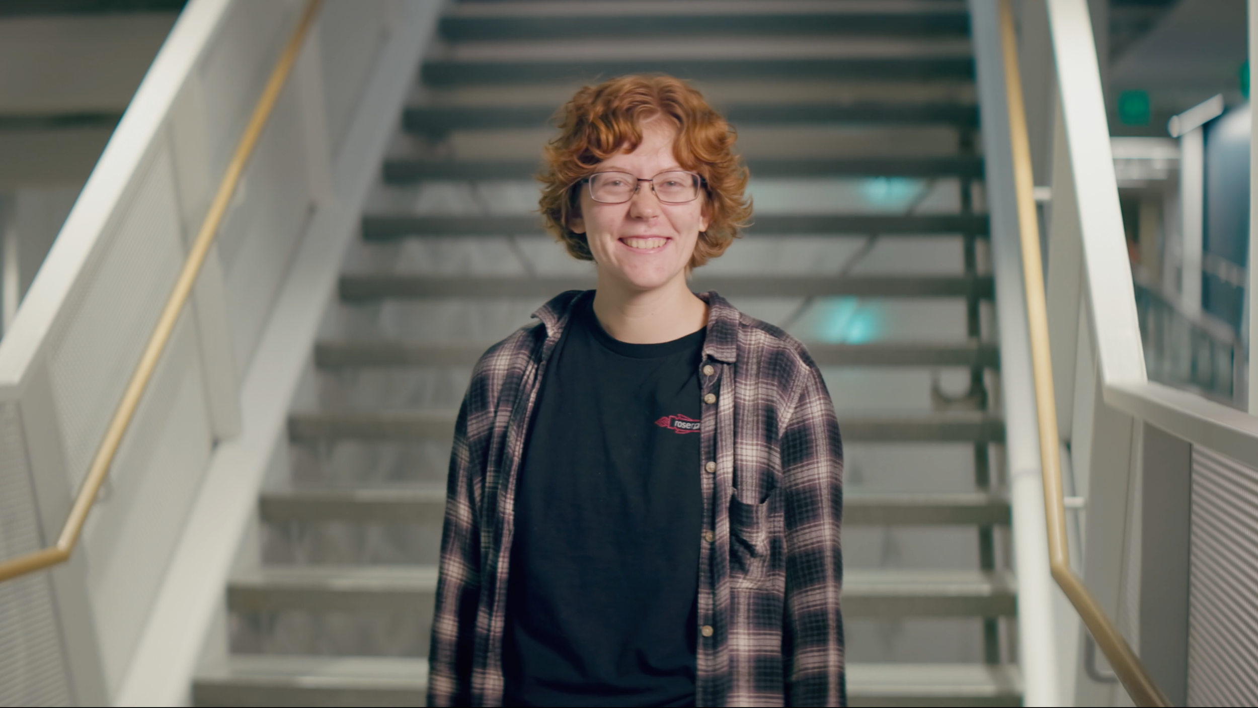 A young person with red curly hair, glasses, and a plaid shirt standing on a staircase in an indoor setting.