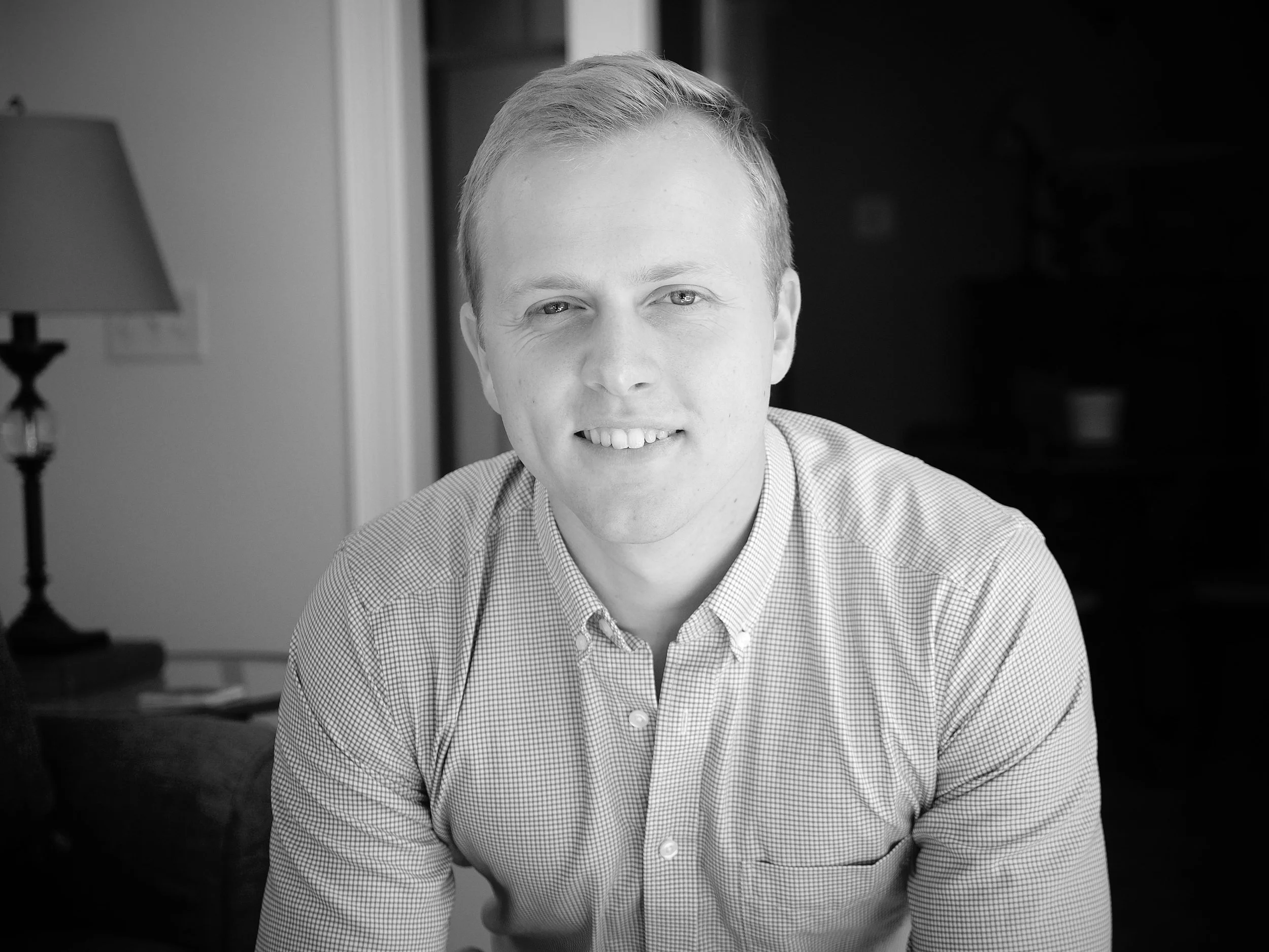 A black and white photo of a smiling young man in a checkered shirt, sitting indoors near a lamp and a door frame.