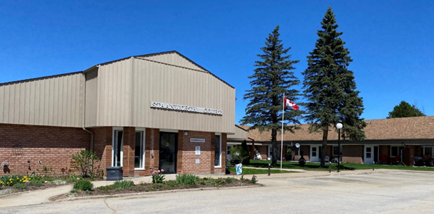 Front view of a small building with a sign reading 'Staanworth Court,' flanked by two tall pine trees and a flagpole with the Canadian flag, in a suburban area with a parking lot and a row of house-like buildings.