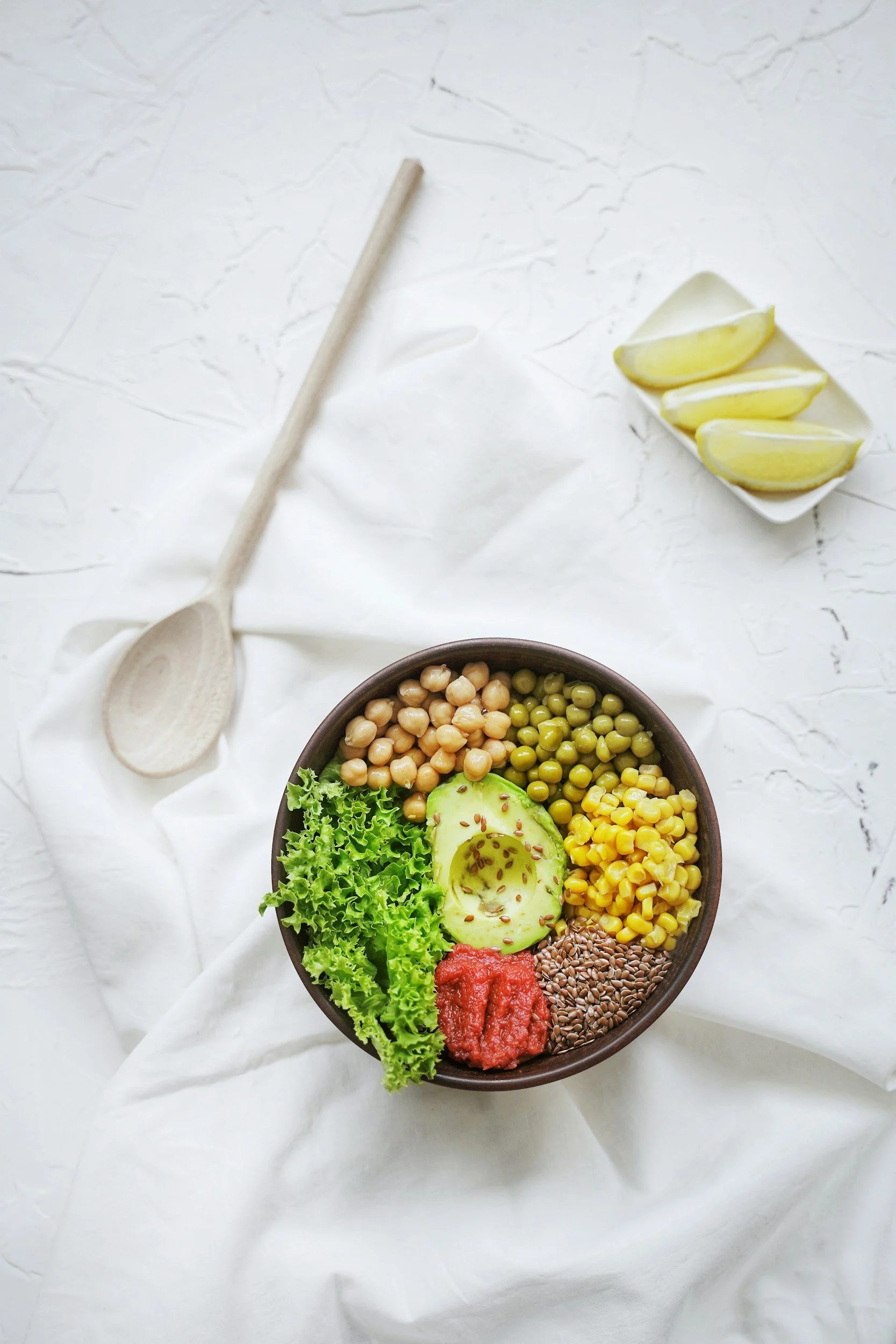 A bowl of a healthy salad with lettuce, avocado, corn, tomato, flaxseeds, and hummus, placed on a white cloth with lemon wedges, a wooden spoon, and a bowl of chickpeas and peas on the side.