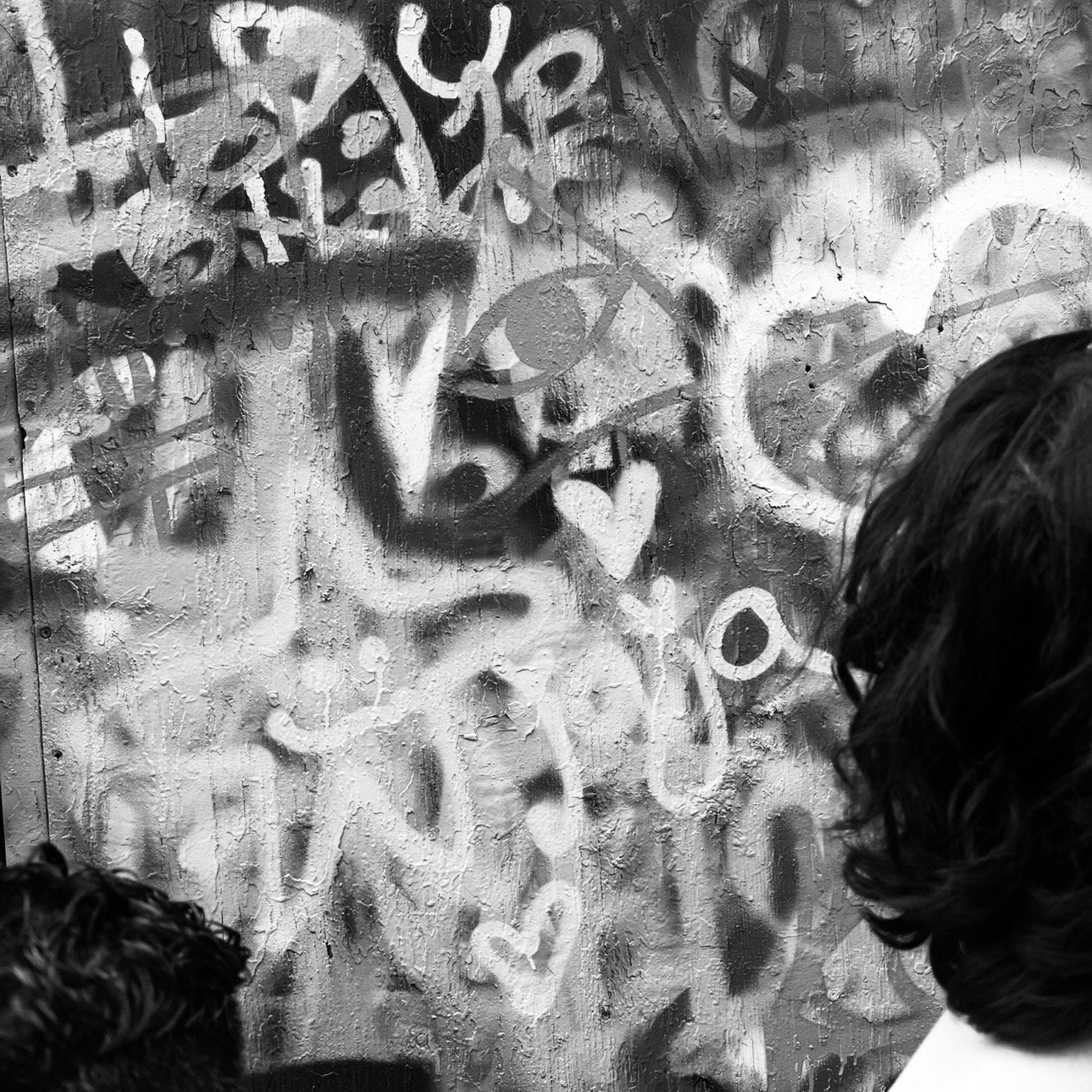 Black and white photograph of a textured wall with graffiti art. The graffiti includes various symbols such as a peace sign, a heart, and an eye, along with other abstract markings. Two people with dark hair are partially visible in the foreground, likely observing the graffiti.