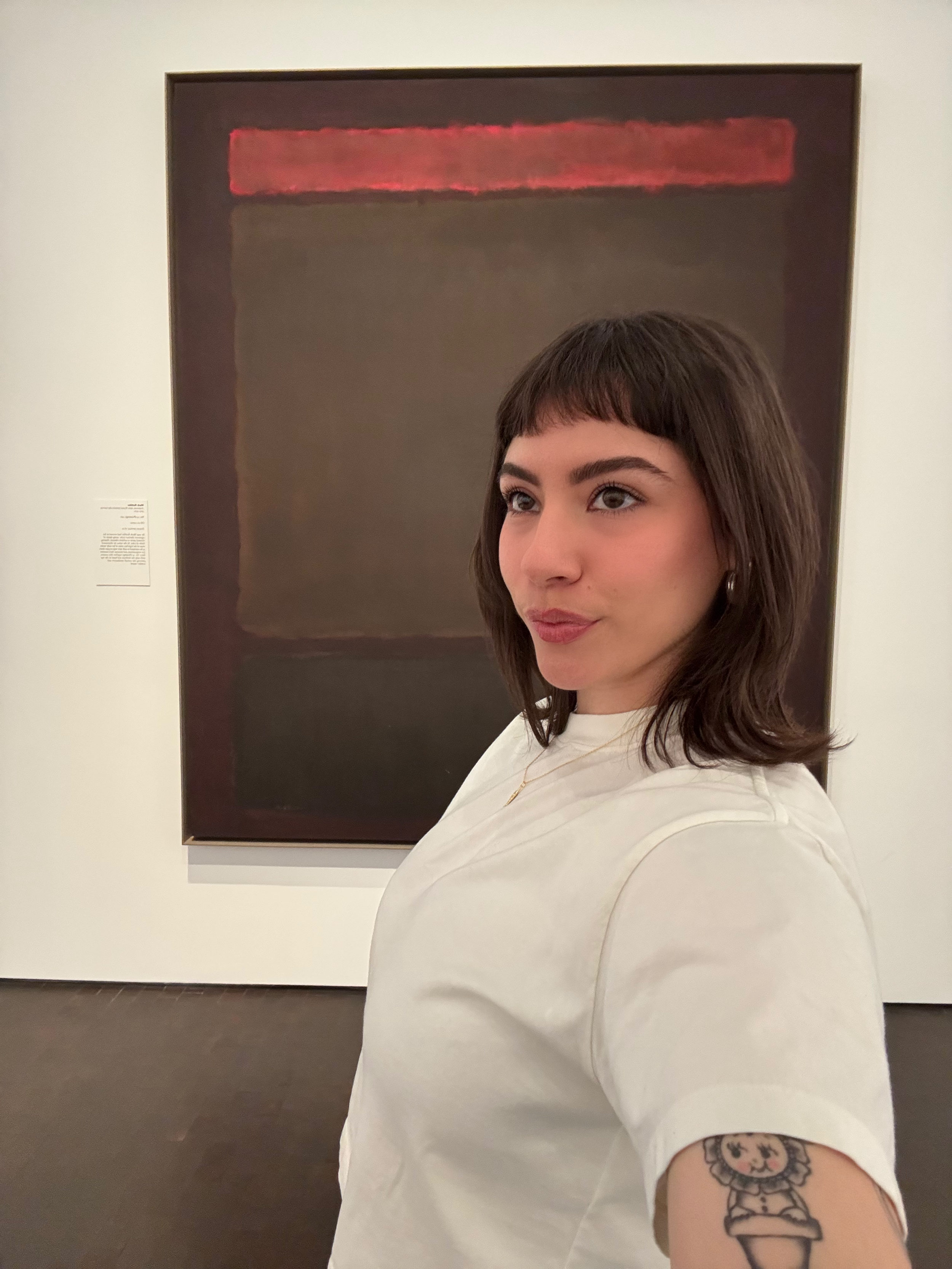 A young woman with dark hair and tattooed arm, wearing a white shirt, standing in art gallery with framed painting on wall behind her.