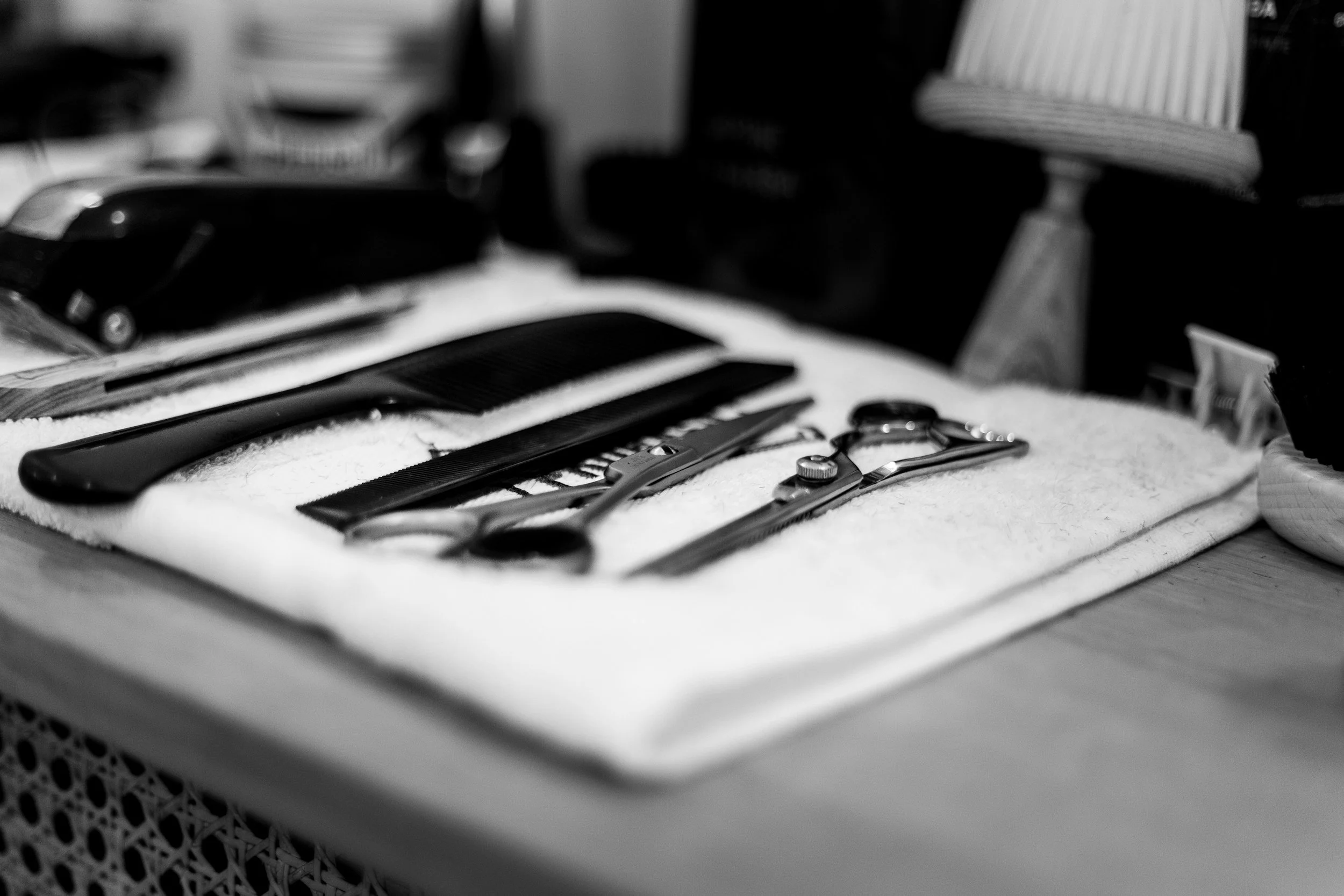 Schwarze Haarschneideschere, Messer und Kämme liegen auf einem weißen Handtuch. Im Hintergrund sind weitere Gegenstände in einem Raum zu sehen.