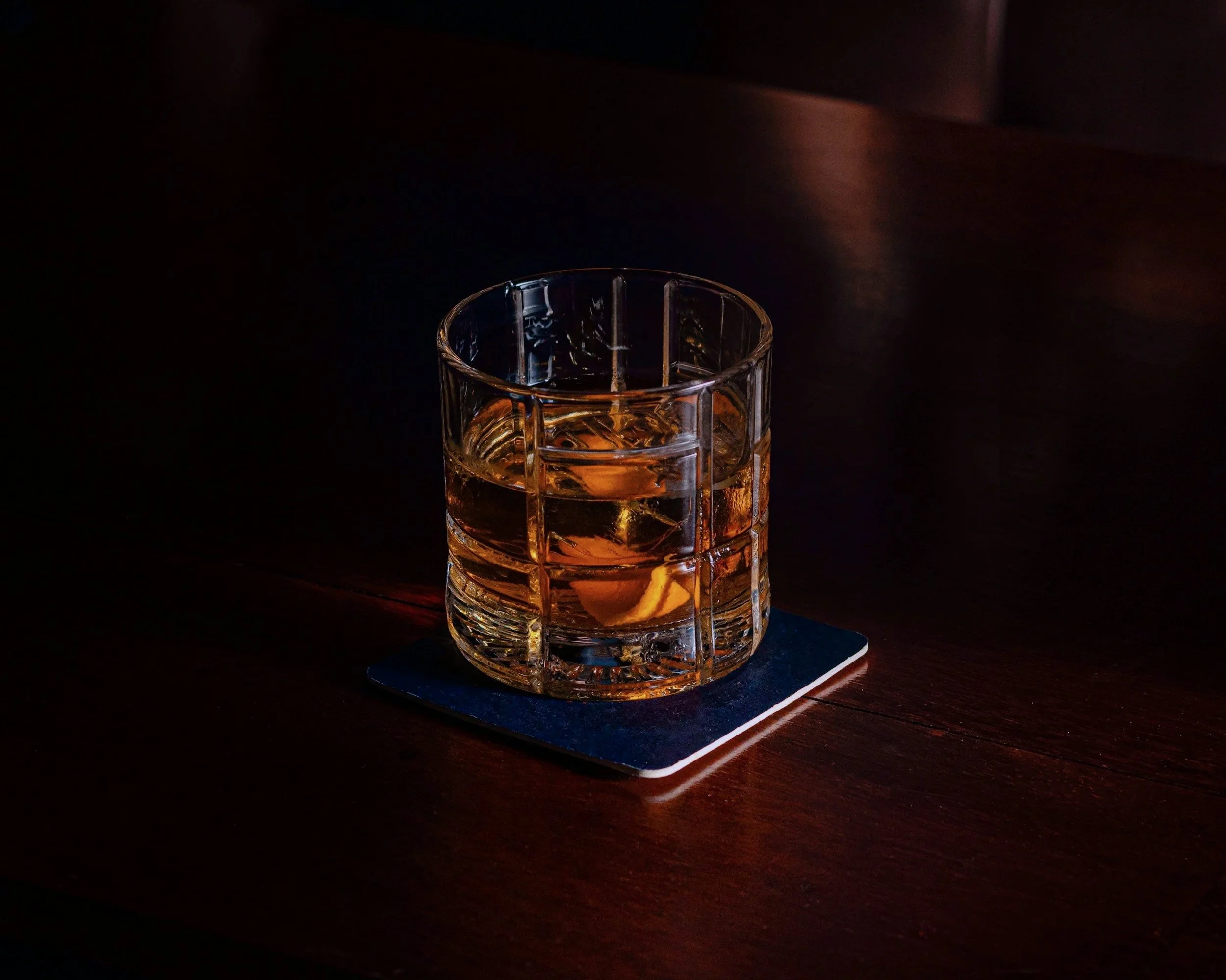 Ein Glas mit Whiskey auf einem dunklen Holztisch, auf einem schwarzen Untersetzer.