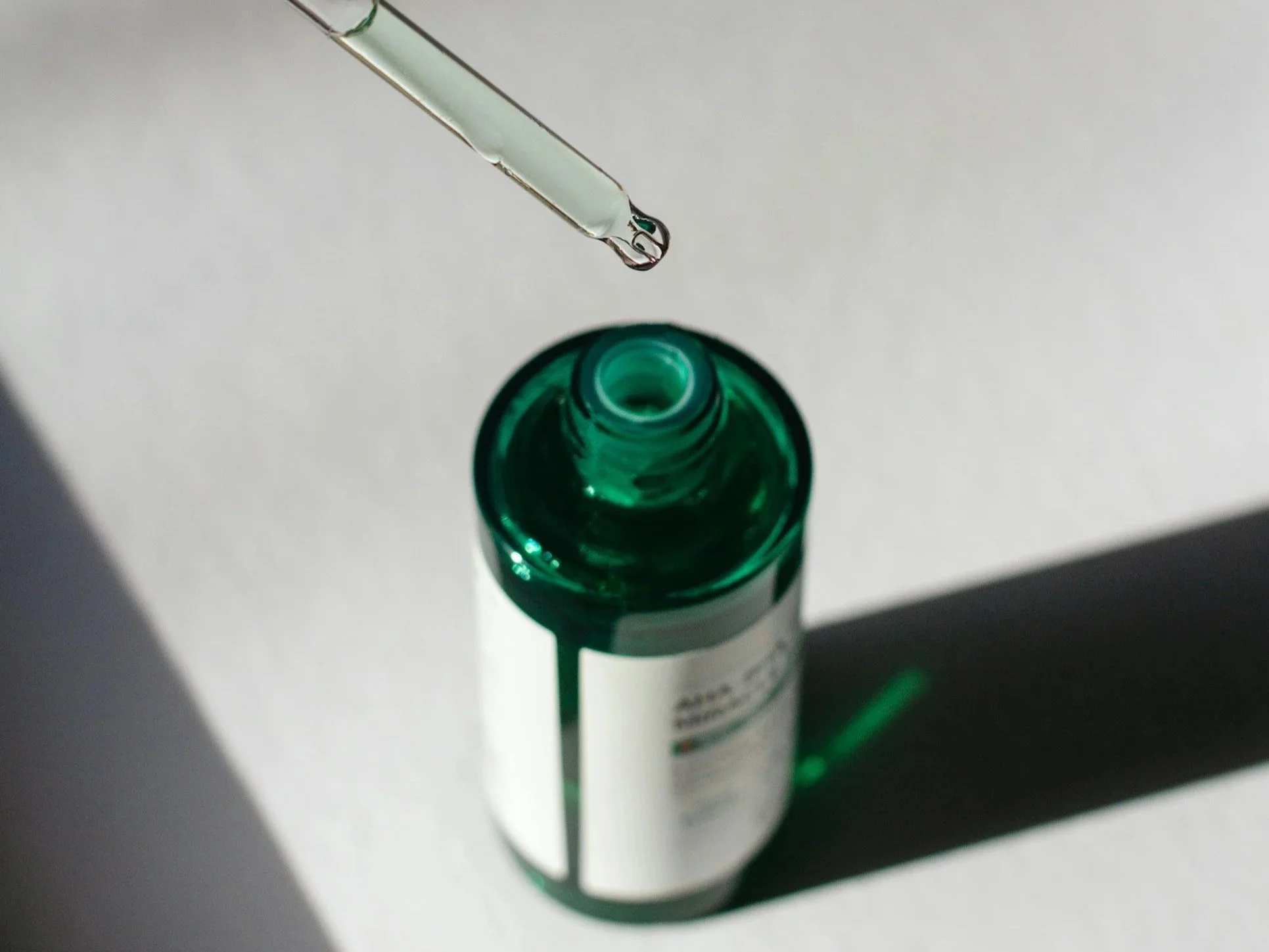 Ein Tropfen Flüssigkeit erscheint an der Spitze einer Pipette über einer grünen Flasche auf weißem Hintergrund.