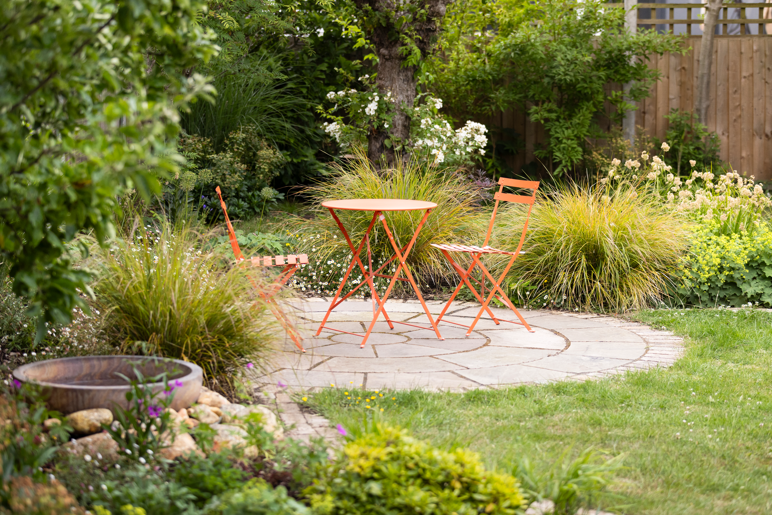 A small patio with a round orange metal table and two matching orange chairs surrounded by lush green plants, trees, and flowers in a backyard garden.