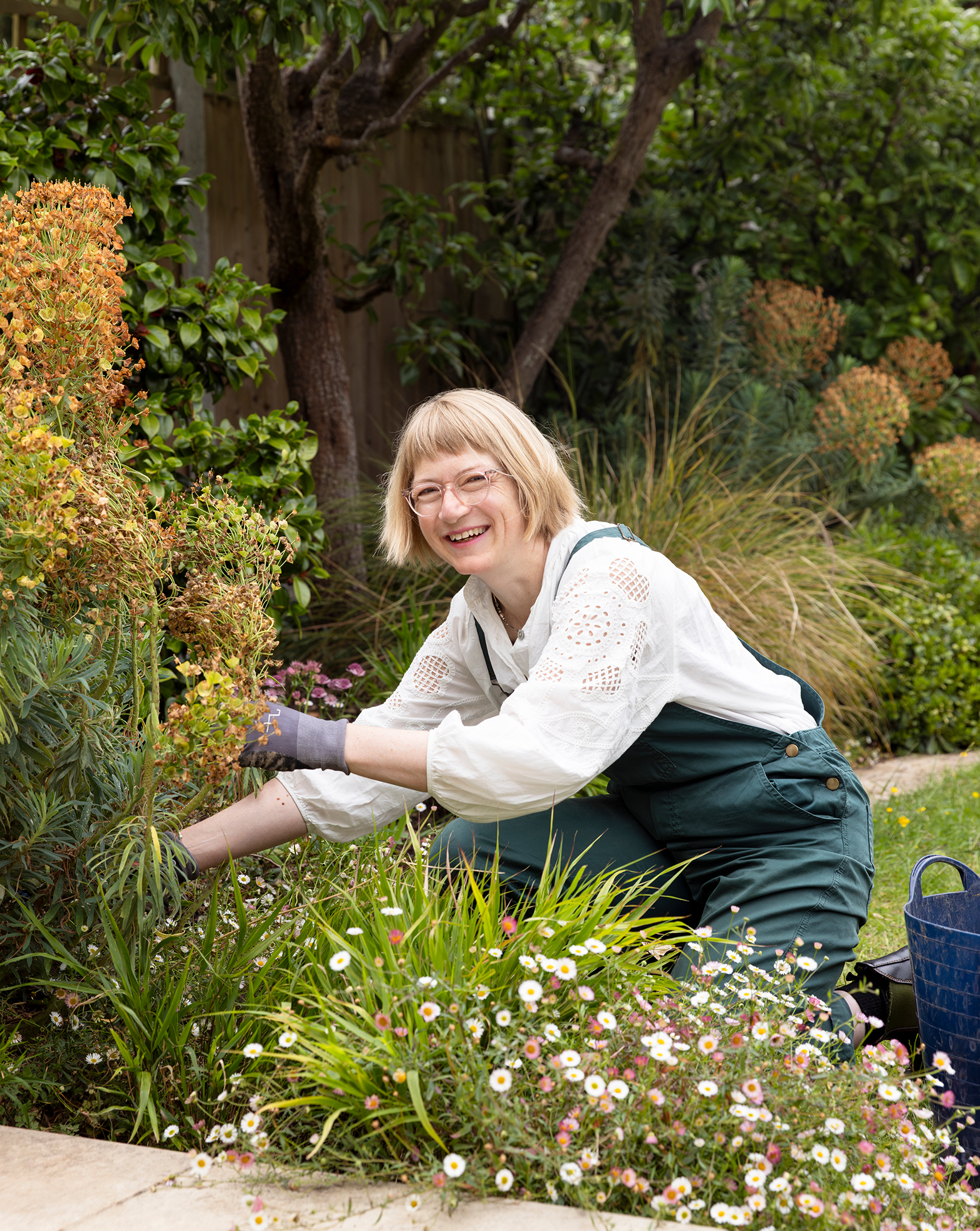 A woman with blonde hair, glasses, and a white blouse gardening among white daisies and various other plants in a lush backyard with trees and a wooden fence.
