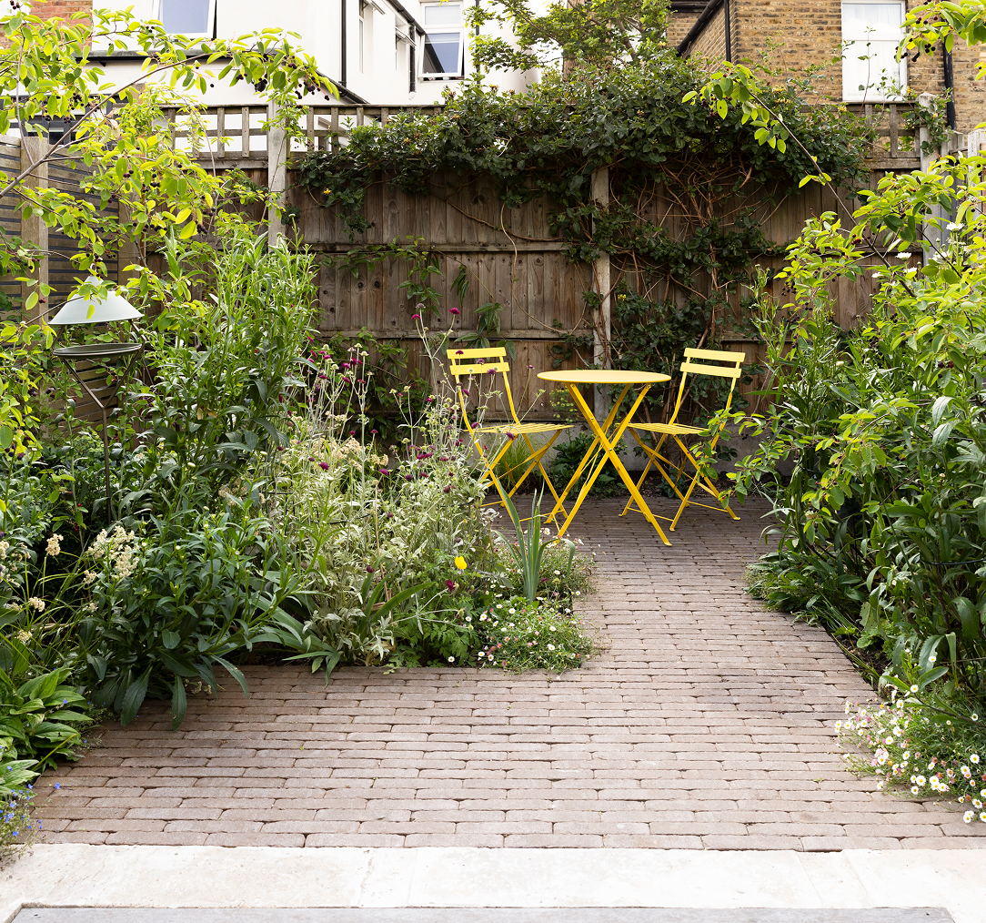 A cozy urban garden with a brick pathway, lush green plants, and a small yellow outdoor table with matching chairs, enclosed by a wooden fence and neighboring brick buildings.