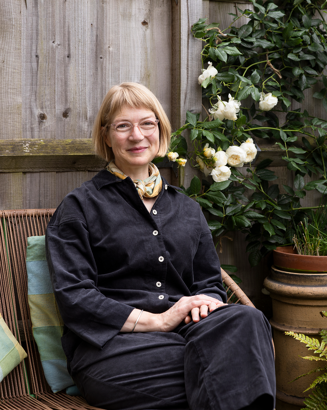 Lucy Pearson with blonde hair and glasses sitting on a wicker outdoor chair with pillows, in a garden setting with plants and white flowers in the background.