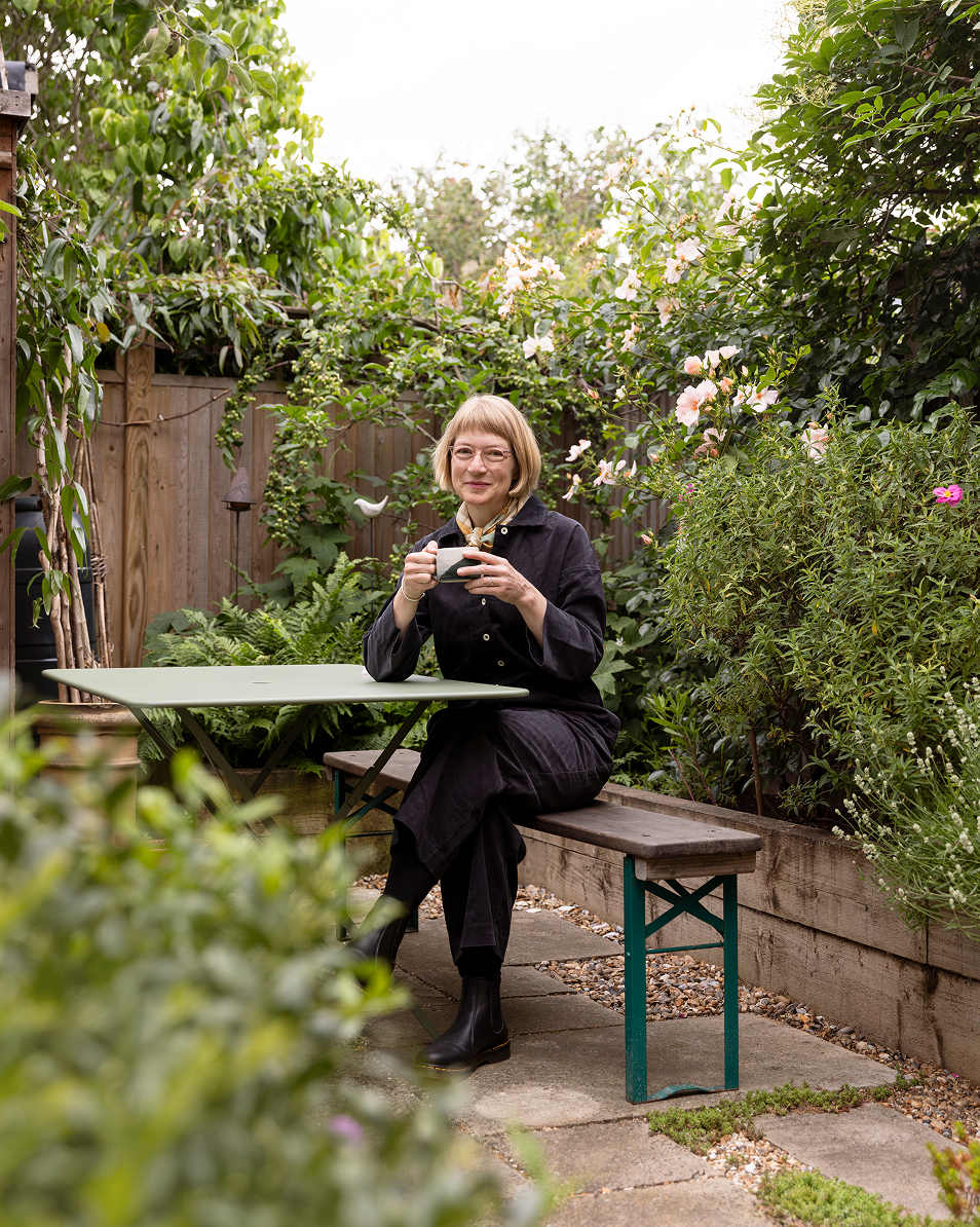 A woman with blonde hair and glasses sitting at a small table in a garden, holding a mug, surrounded by lush green plants and pink flowers.