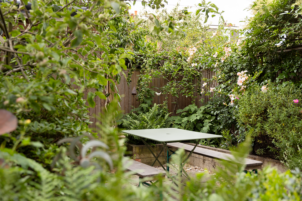 A lush garden with dense green foliage and blooming pink and white flowers surrounding a small outdoor table with benches.