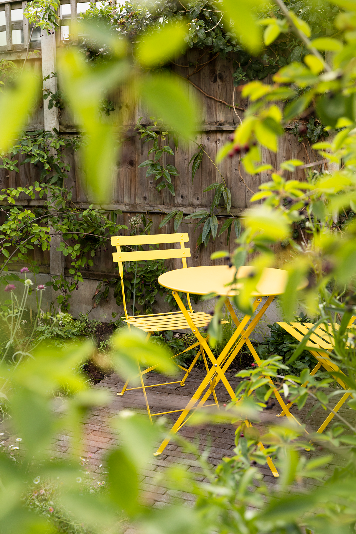 Bright yellow outdoor patio furniture set with a table and two chairs on a brick patio, surrounded by green plants and a wooden fence.