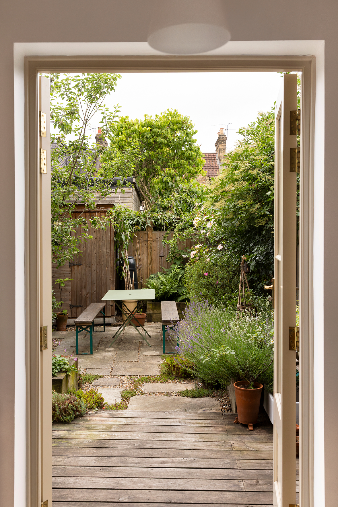 View from open French doors into a small, lush garden with a wooden fence, potted plants, a small table with benches, leafy trees, and flowering plants.