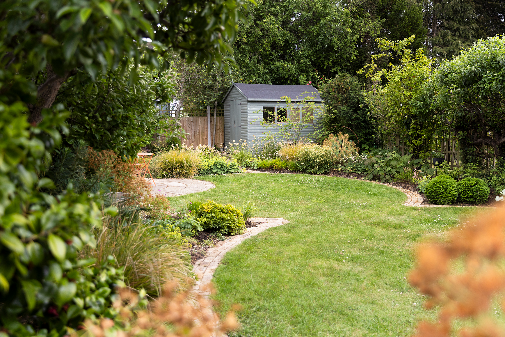A backyard garden with a well-maintained lawn, a curved brick pathway, lush shrubs, trees, and a small gray shed in the background.