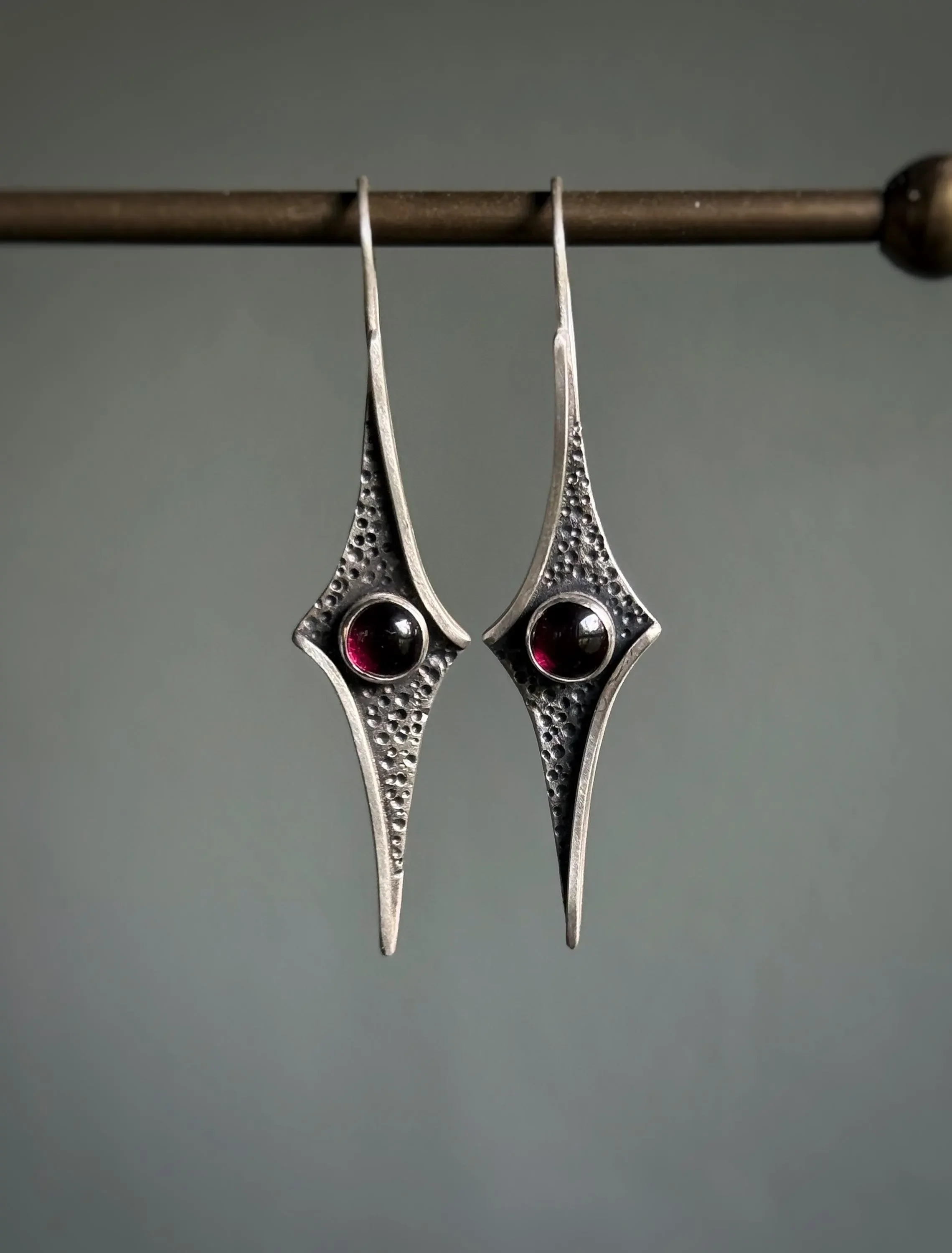 A pair of silver earrings with a textured surface and a deep red gemstone in the center, hanging from a metal rod.