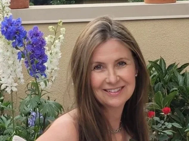 A smiling woman with long brown hair posing in front of a garden with purple, white, and red flowers.