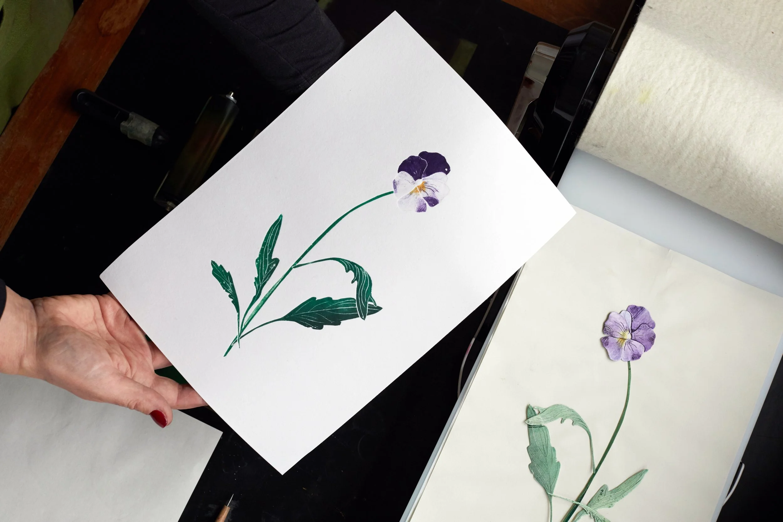 A person's hand holding a white card with a pressed purple and white flower illustration, on a black surface with art supplies nearby.