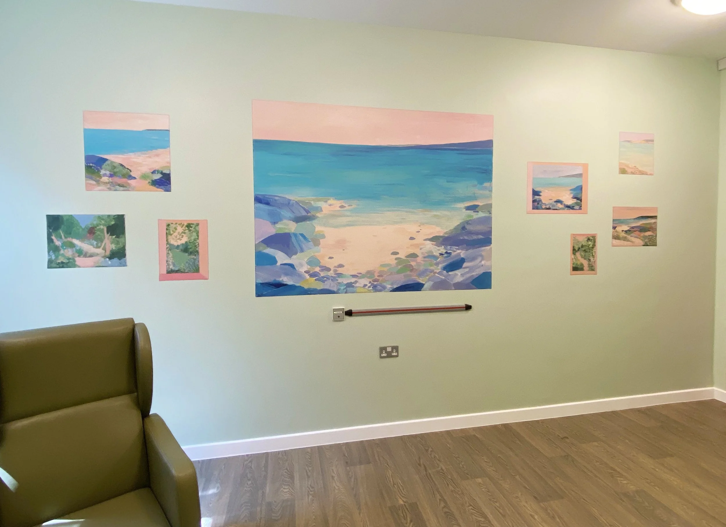A wall decorated with several landscape paintings, including a large painting of a beach scene with rocks and the sea, and smaller paintings of similar coastal scenery. Part of a green upholstered chair is visible in the lower left corner. The floor is wooden, and there are electrical outlets on the wall.