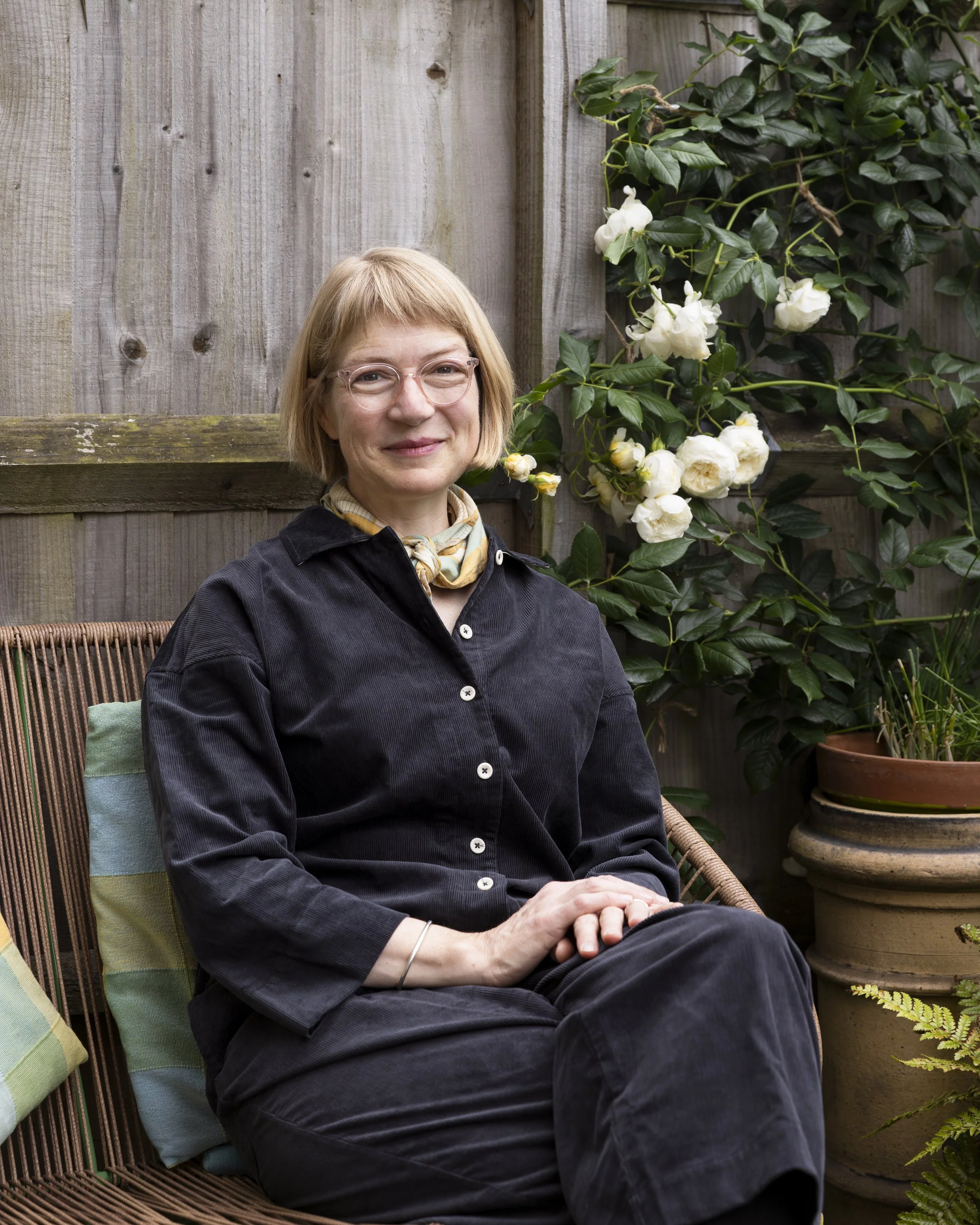A woman with blonde hair and glasses, wearing a black shirt and scarf, sitting on a patio with a wooden fence and green plants and white flowers in the background.