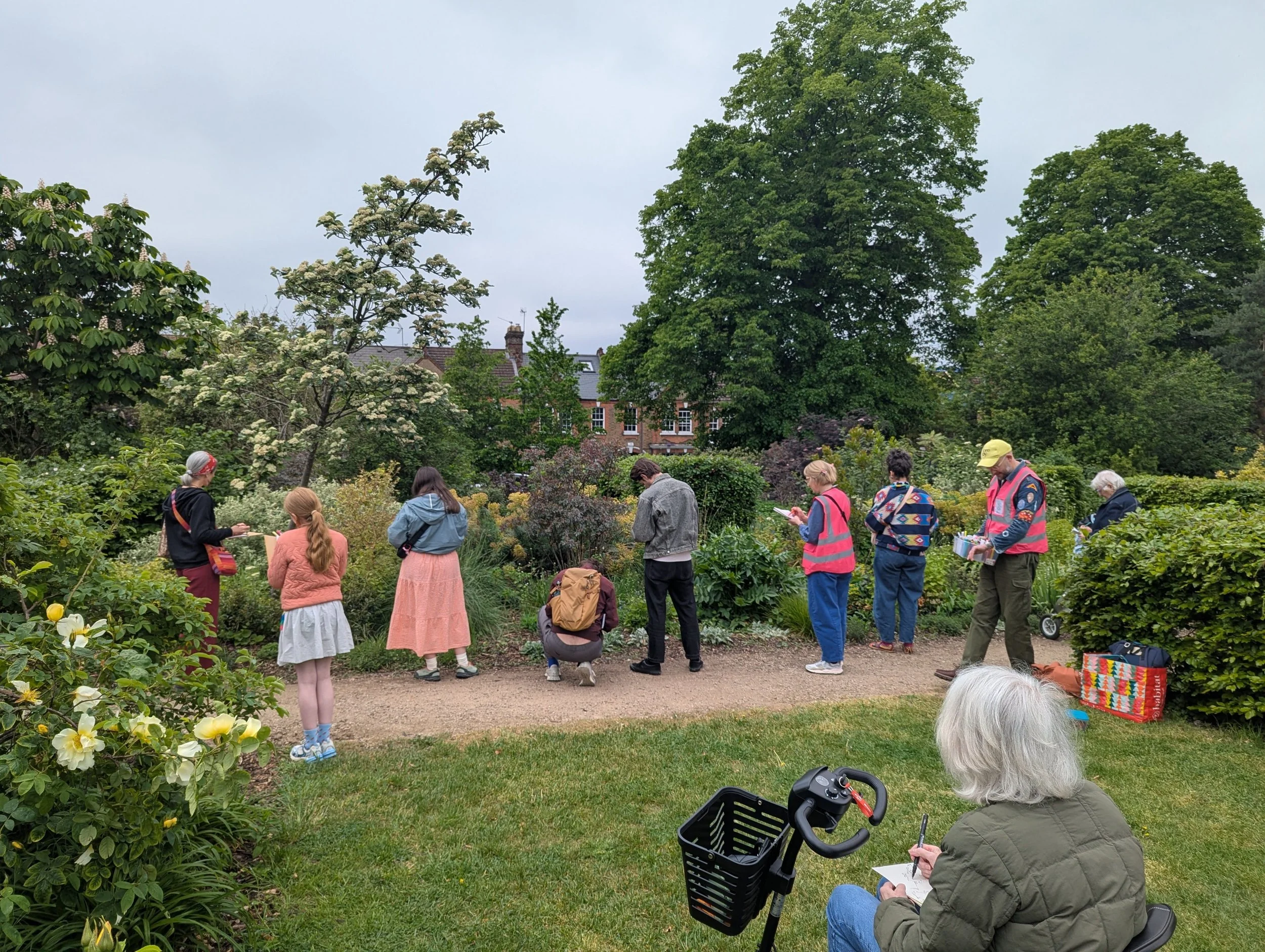 group of people in a garden with lush green trees and buildings in the background, some standing and reading, one person sitting on a scooter writing in a notebook, others engaging in conversation or looking at plants.