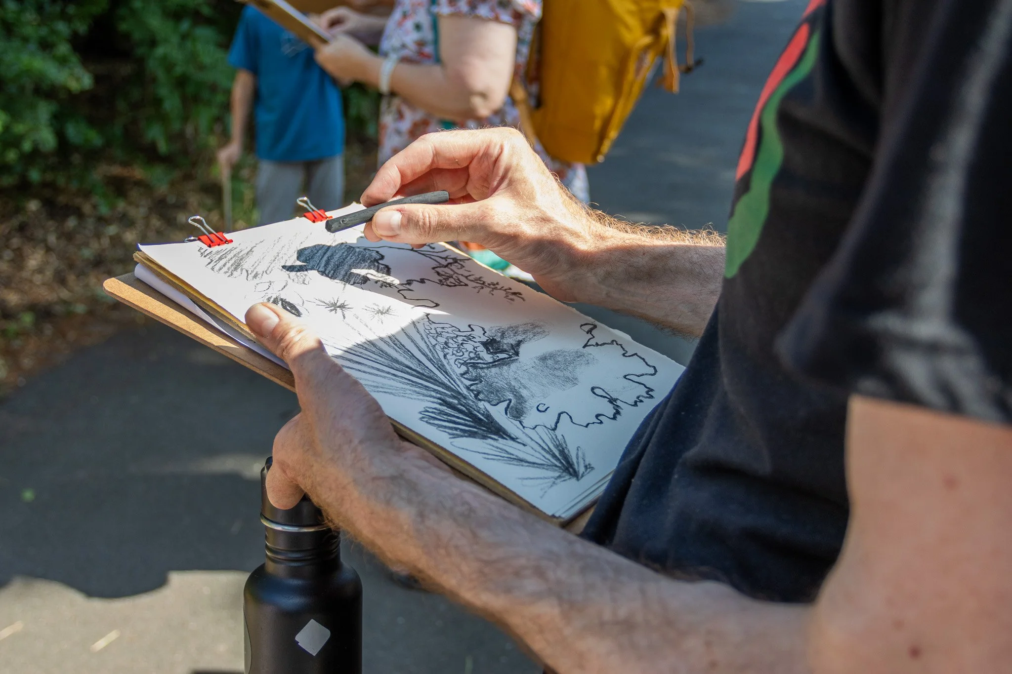 Person holding a clipboard with black and white sketches, outside, with other people in the background.