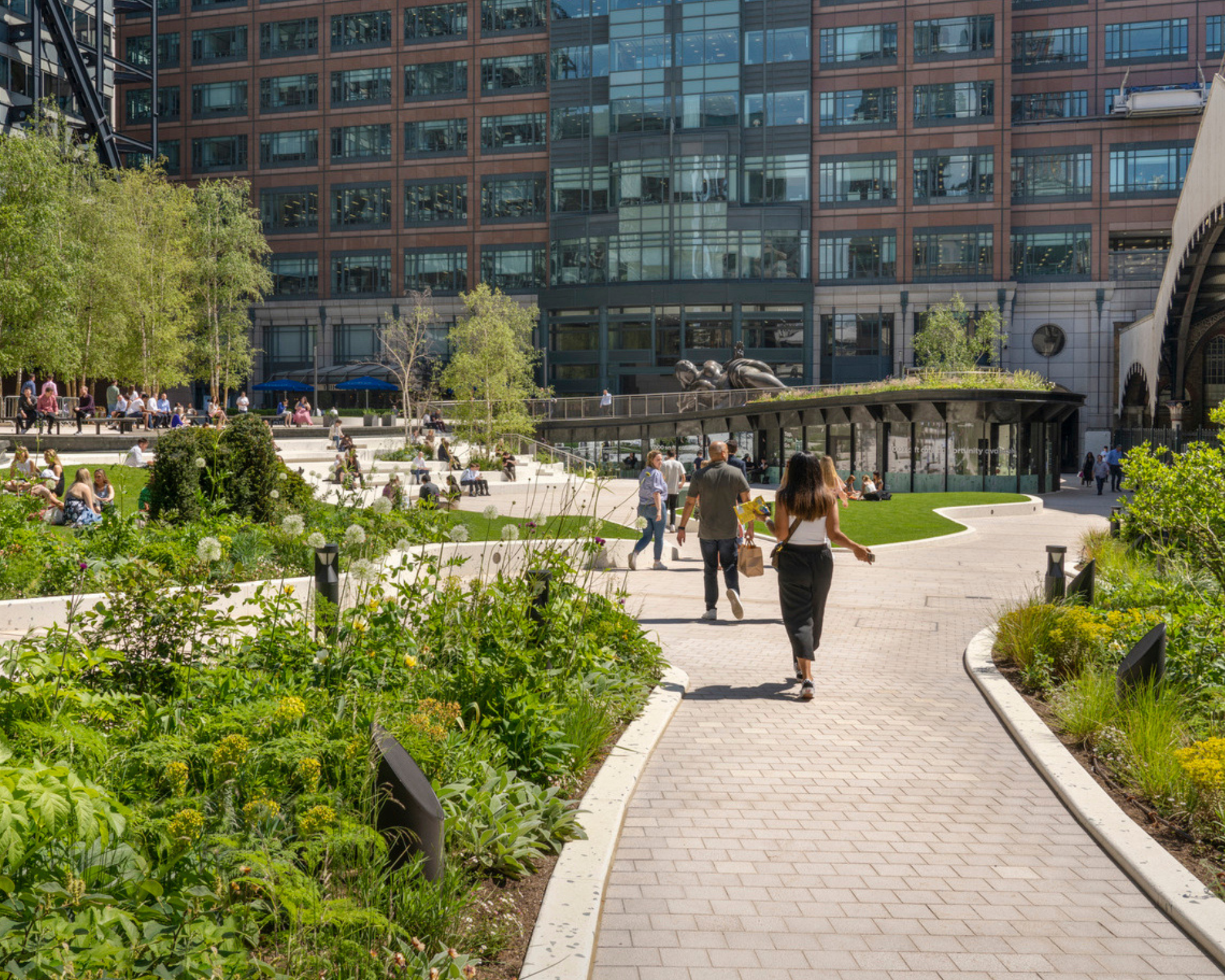 Urban park with greenery, trees, and flower beds in front of a modern office building with glass windows and brick facade, people walking and sitting on benches.
