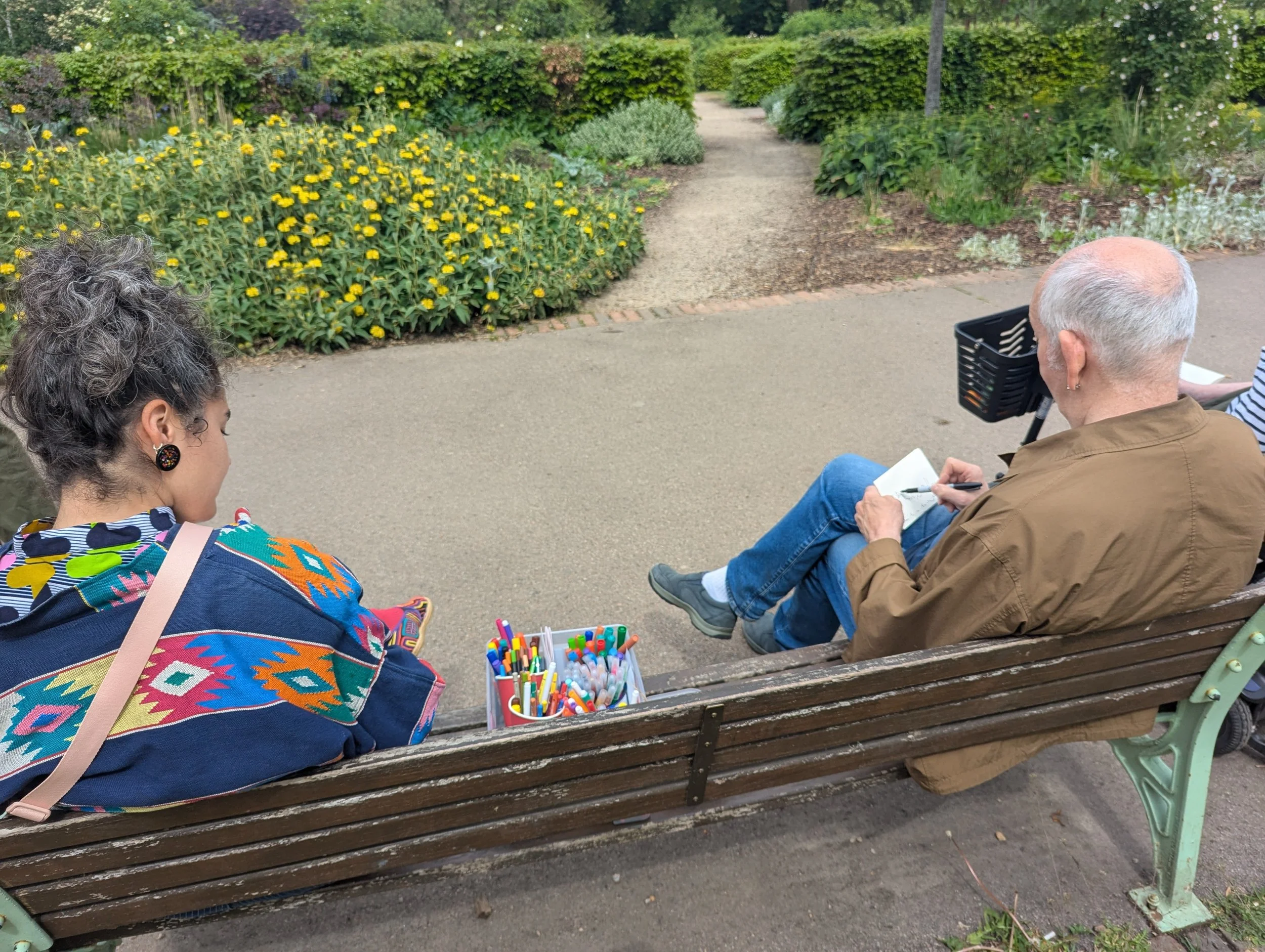 A woman and an older man sitting on a park bench, with a garden path and green bushes in the background. The woman is wearing a colorful patterned top and sitting with her back turned slightly, with a container of colorful pens or markers in front of her on the bench. The man has a bald head with white hair on the sides, and he is writing or drawing in a notepad, with a black basket attached to his bike handlebar nearby.