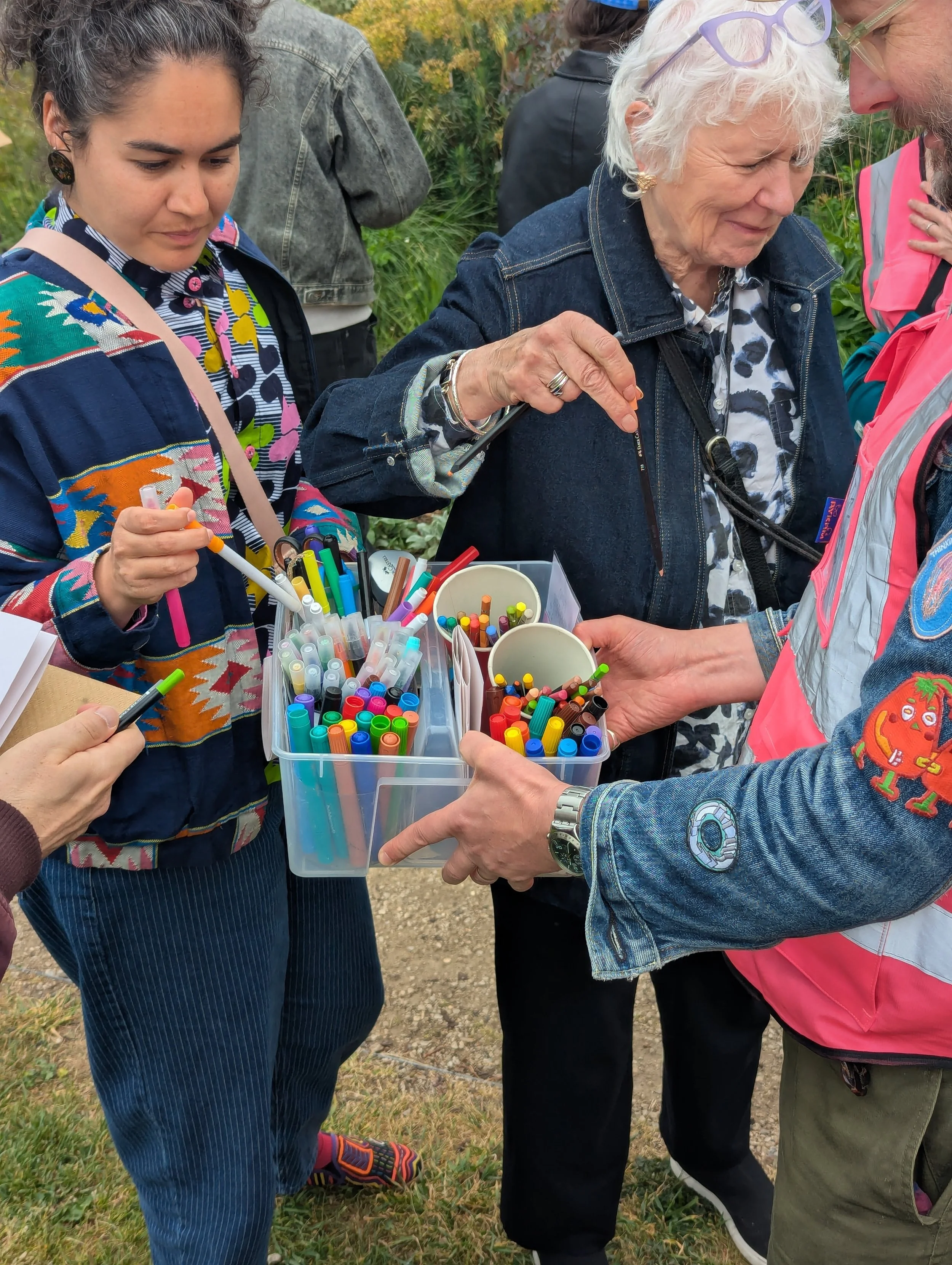 Group of people outdoors examining and sorting colorful markers from containers, with trees and grass in the background.