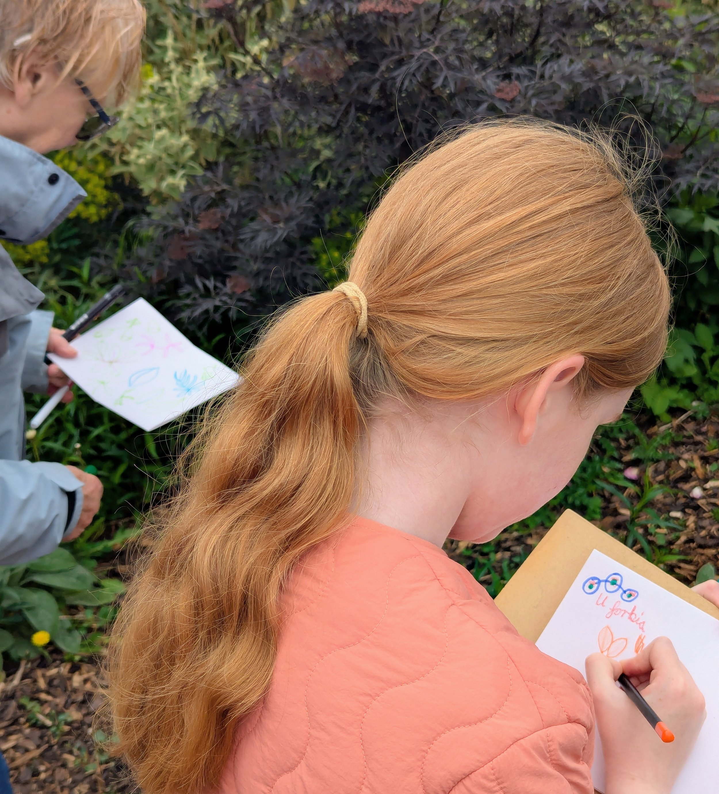 Close-up of a red-haired woman drawing on a piece of paper, with children around her, outdoors with greenery and purple leaves in the background.